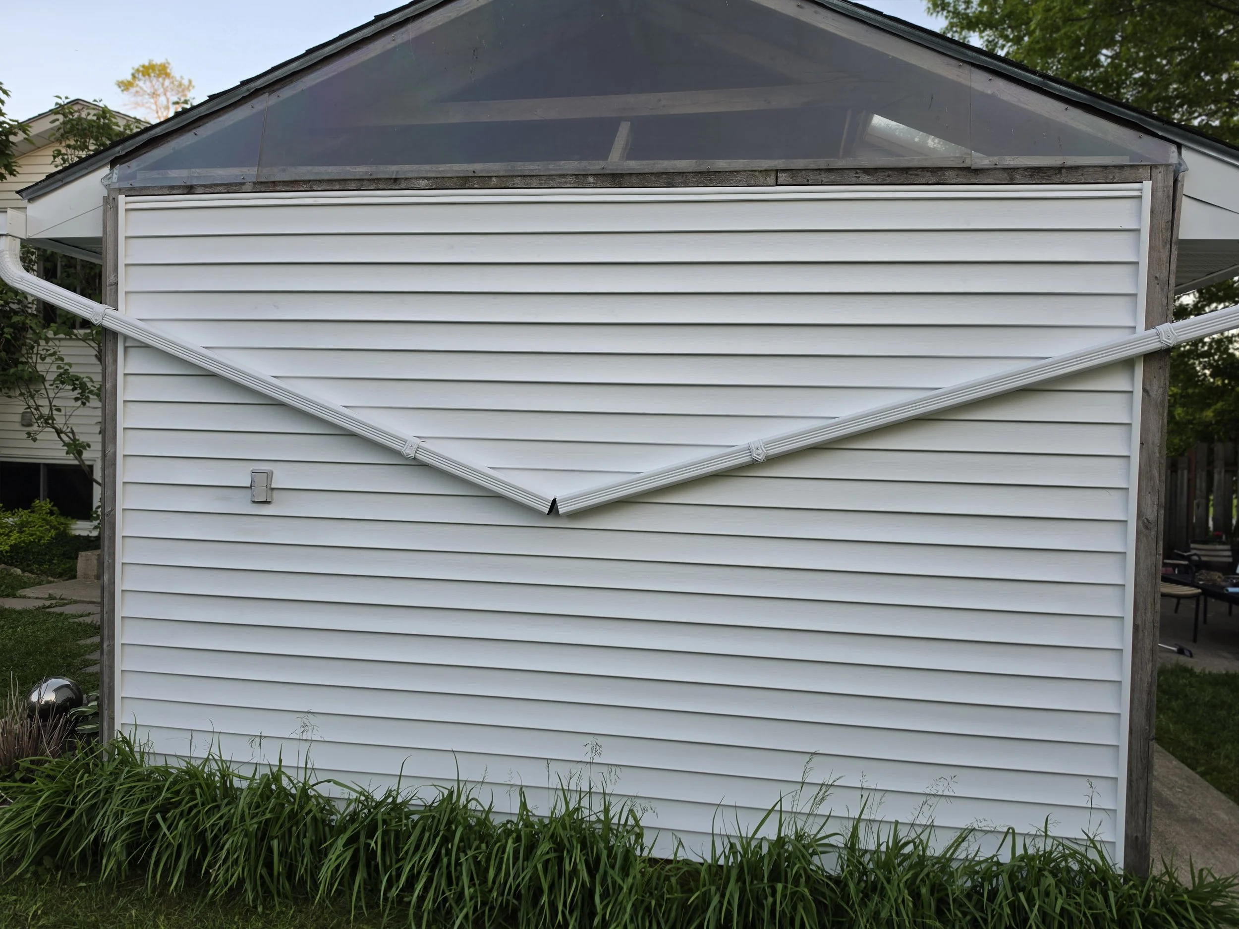 A house with white vinyl siding and a shed with ladders on its exterior, surrounded by green grass and trees.