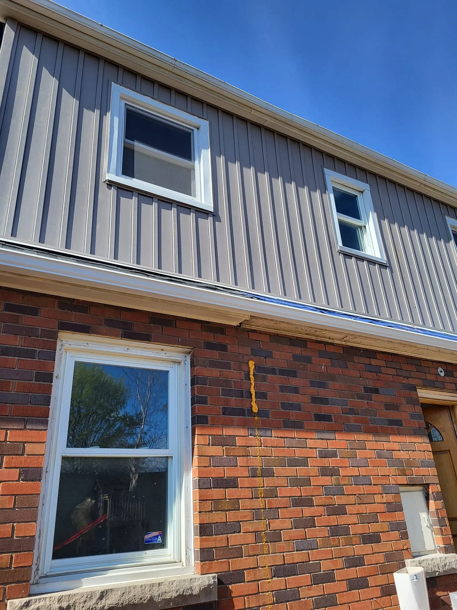 The image shows a residential house exterior with a combination of brick and siding walls. There are two white-framed windows on the second floor and one window on the first floor, with reflections of trees visible in the glass. A yellow string or co