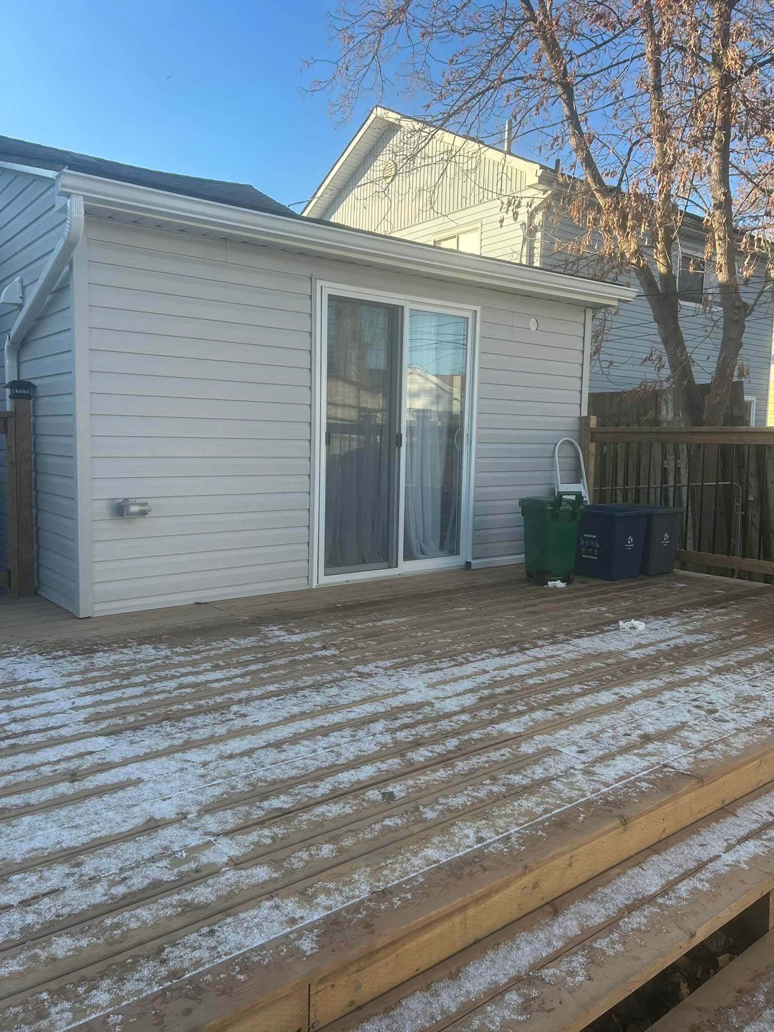 Backyard wooden deck with snow, a small white shed with sliding glass door, green and black trash cans, a wooden fence, and a leafless tree against a clear blue sky.