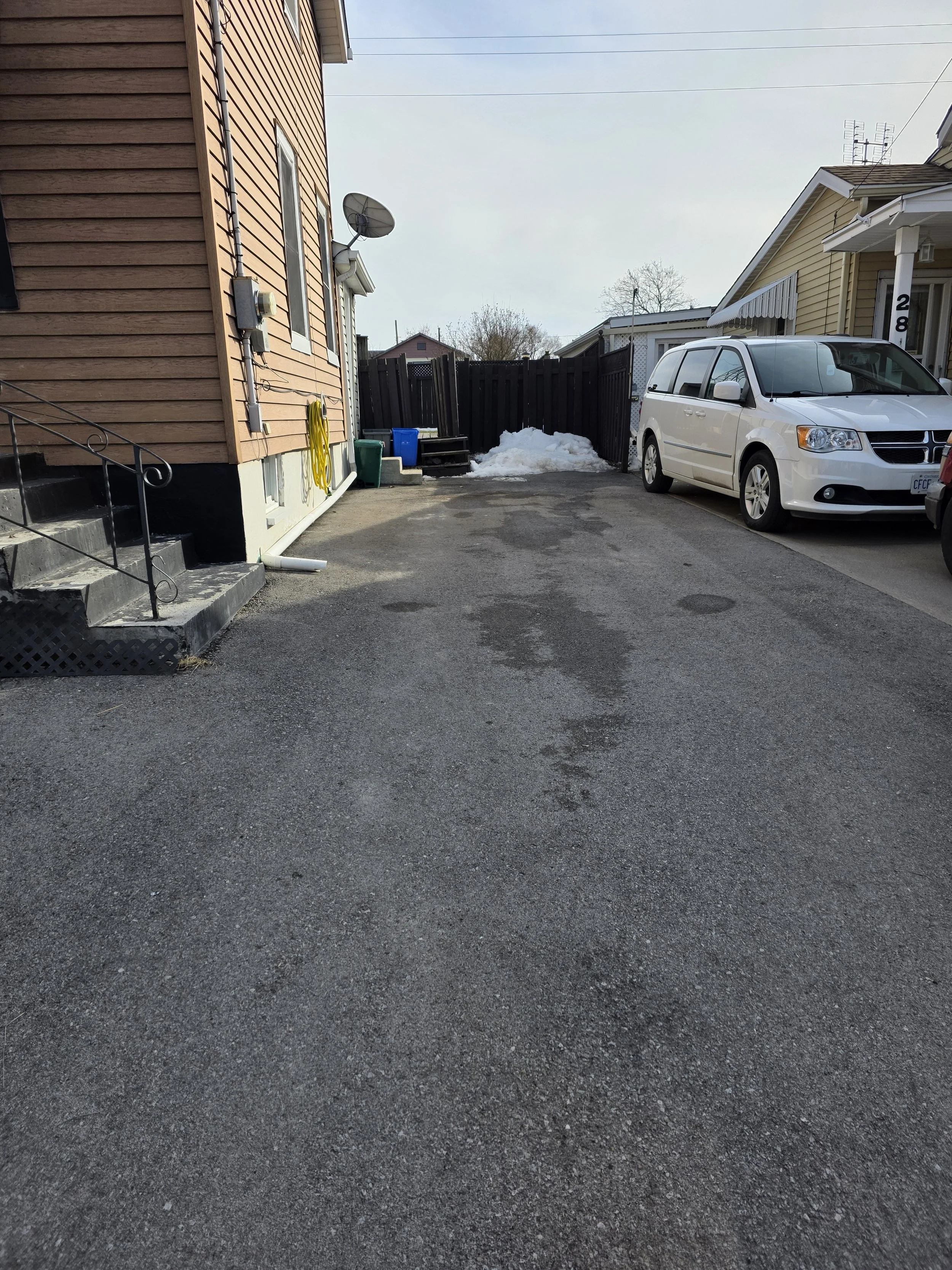 Residential driveway with parked white minivan, snow pile, black fence, and neighboring houses, under a cloudy sky.