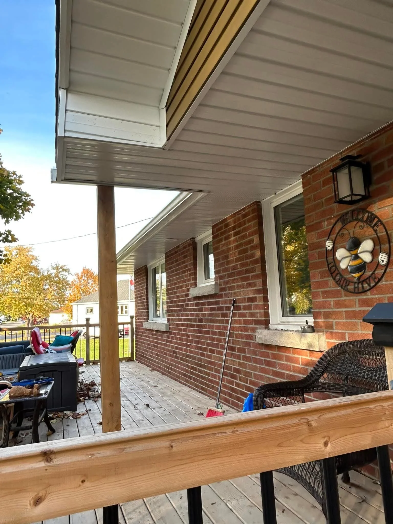 View of a residential porch with wooden flooring, red brick walls, windows, outdoor furniture, a lantern-style light fixture, and trees in the background with autumn foliage.