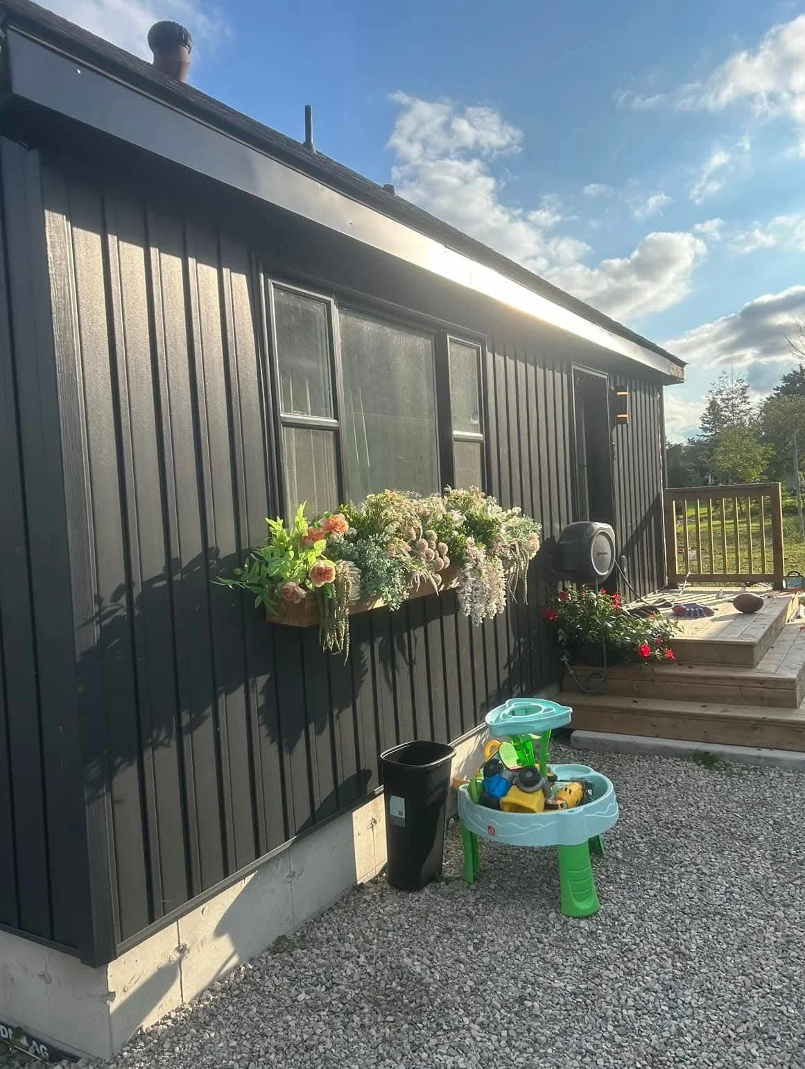 View of a black house exterior with a flower box, a speaker, and a child's toy table with toys, a black trash can, and a deck with outdoor furniture in the background.