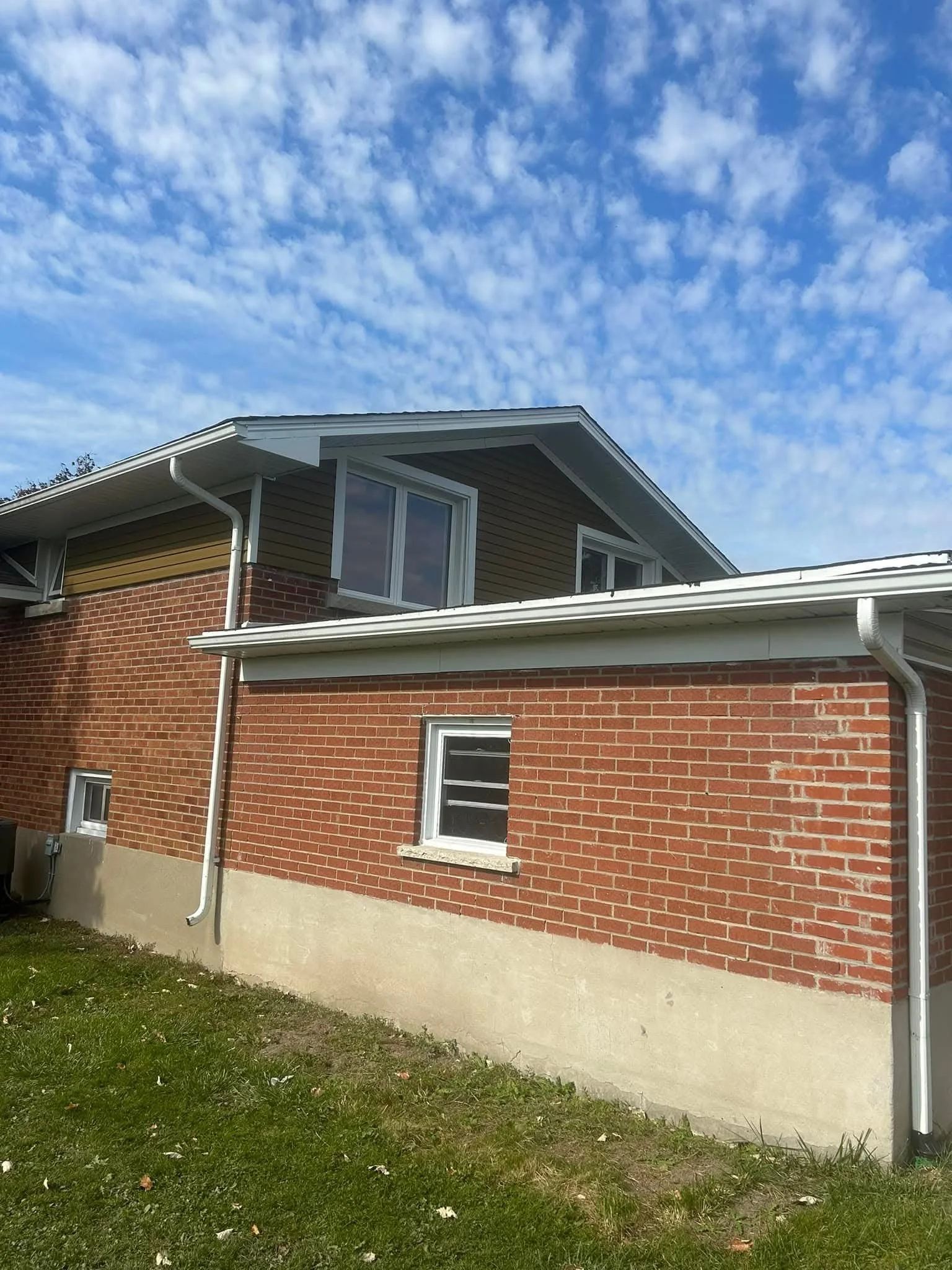 The exterior of a brick house with a sloped roof, two visible windows, and a gutter system, set against a blue sky with clouds and a grassy yard in the foreground.