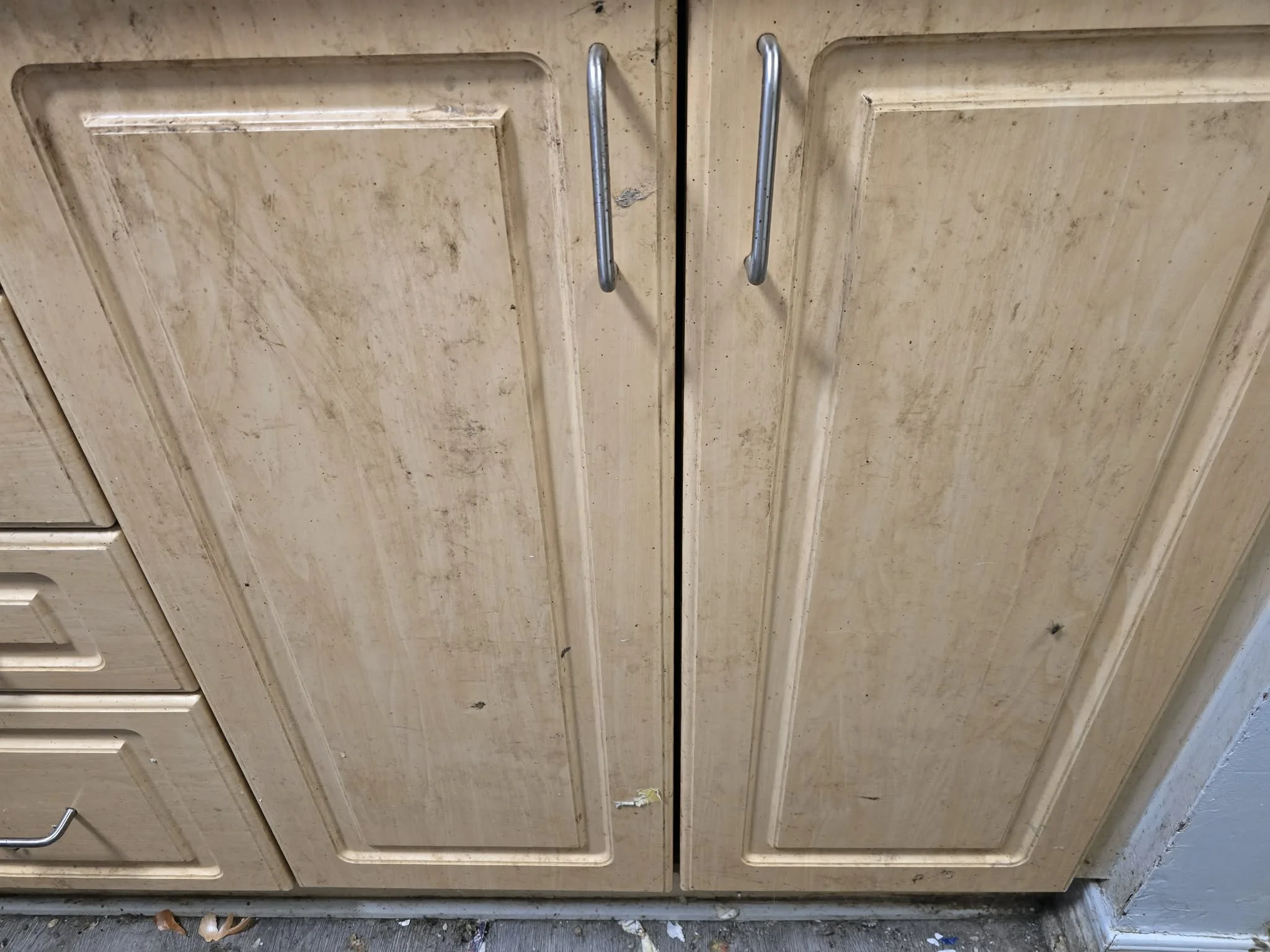 A beige wooden cabinet with two metal handles in the center, showing visible dirt and scratches, situated on a concrete floor with some debris.