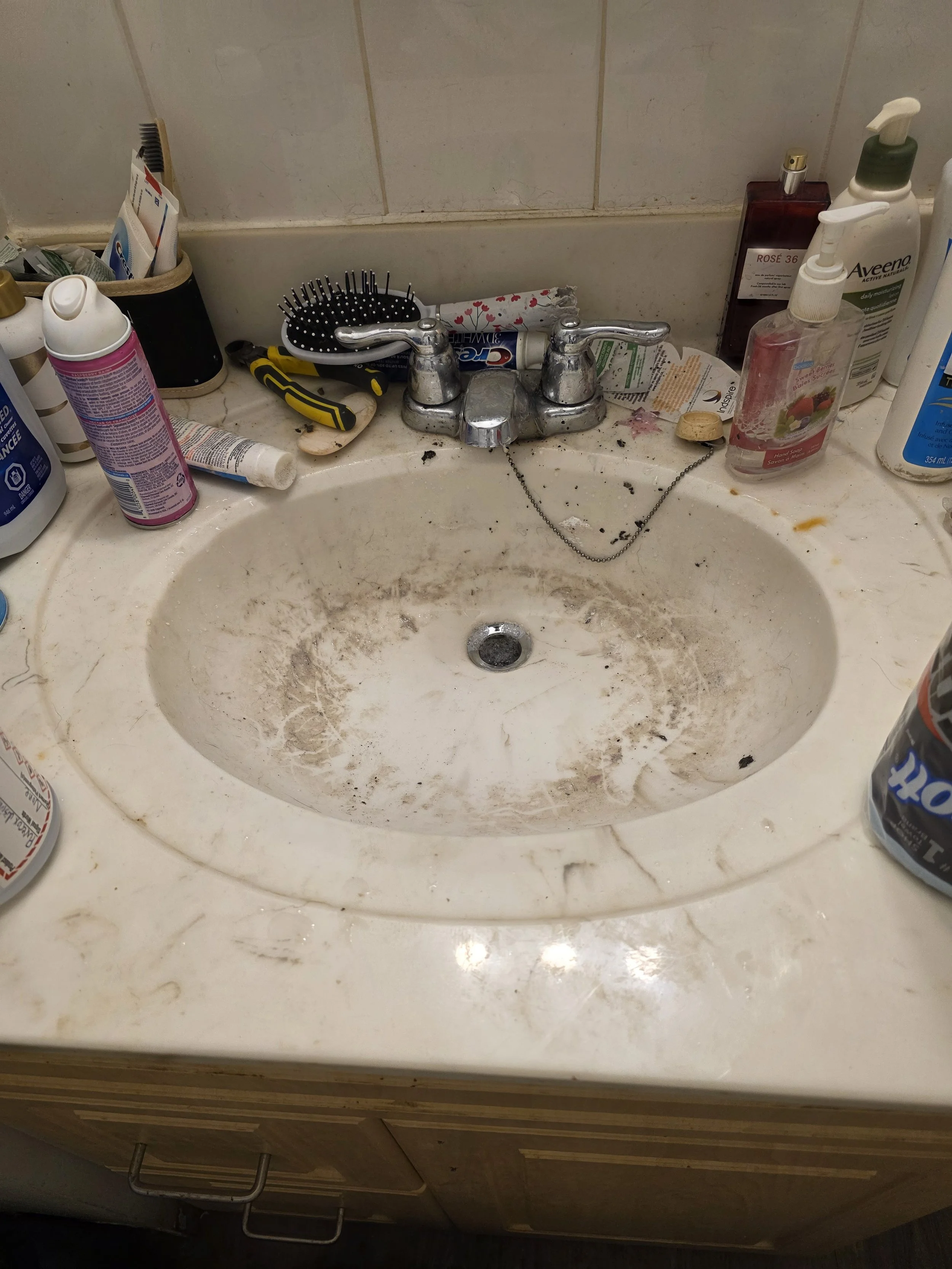 Dirty bathroom sink with dirty countertop, grime, and scattered toiletries including hand soap, lotion, mouthwash, and various tubes and bottles, along with hairbrush and tools, indicating neglect and clutter.