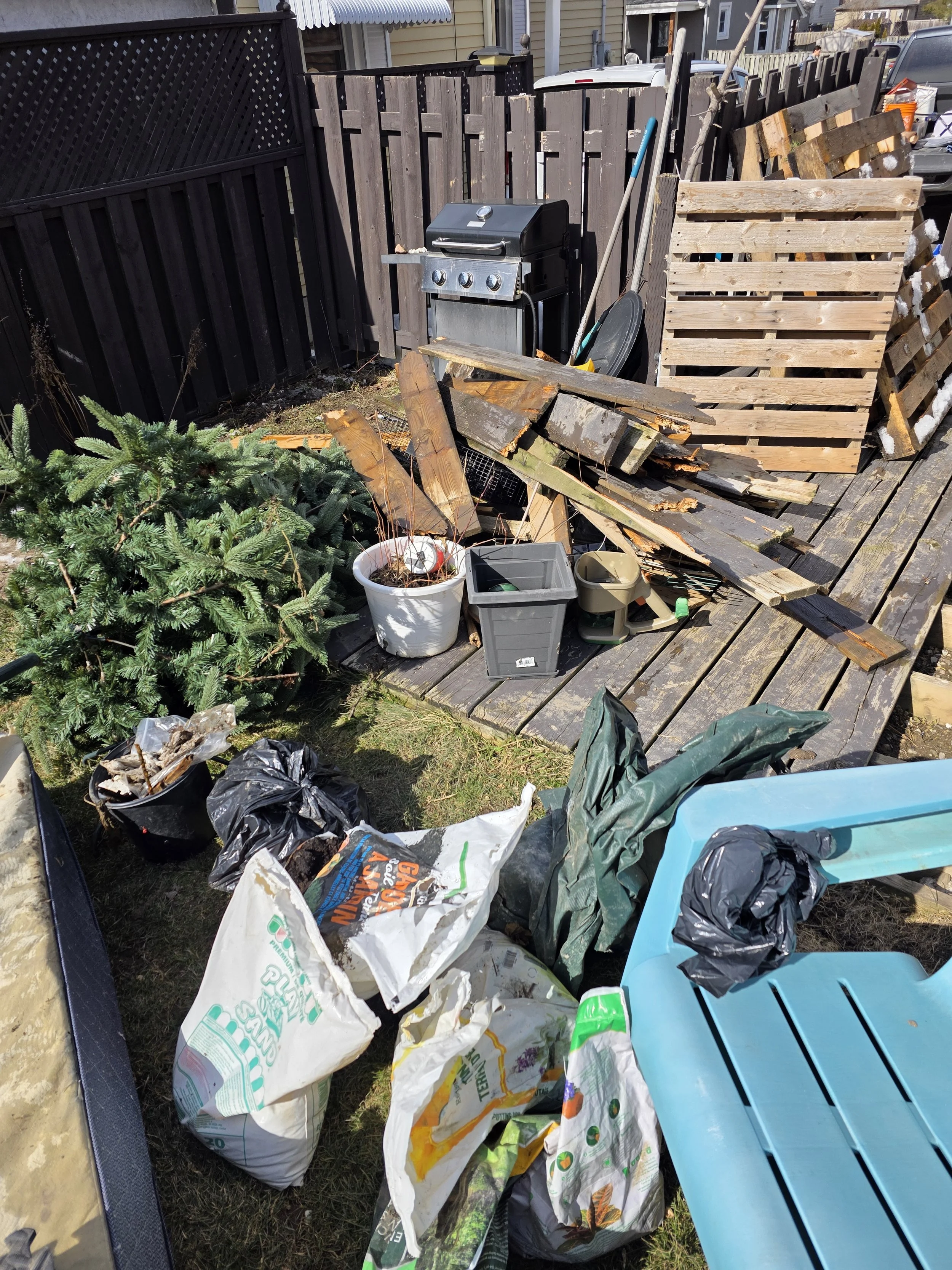 Backyard with a broken wooden deck, scattered trash, a small green pine tree, a white pot, gardening supplies, a black grill, and a blue plastic storage container.