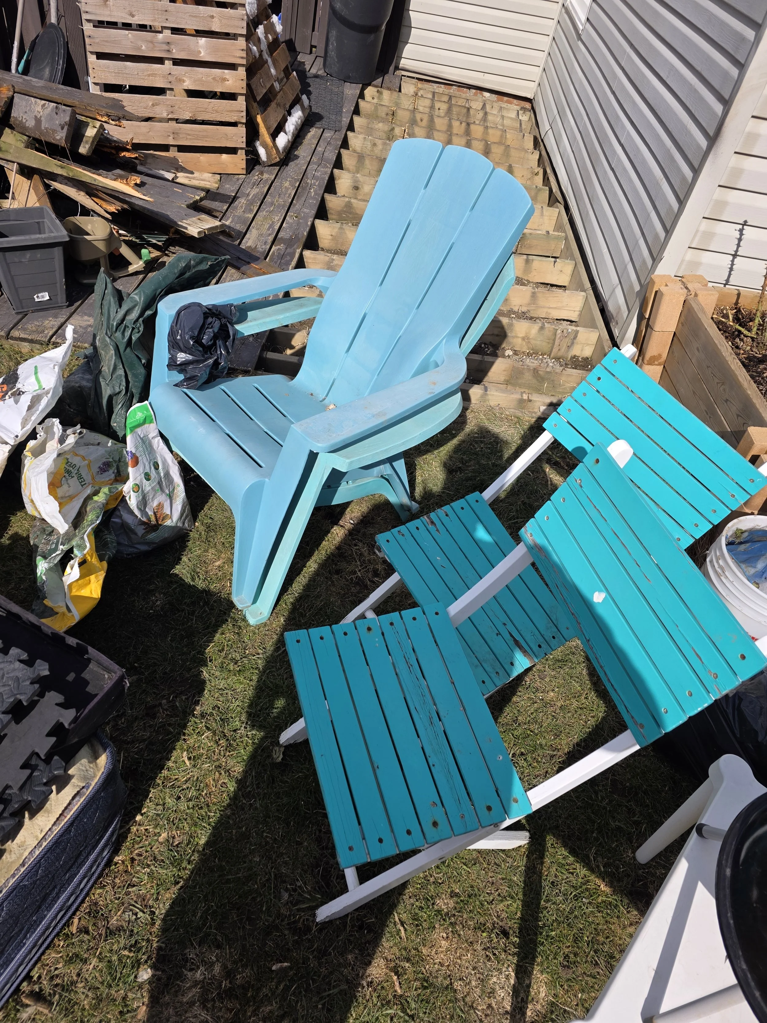 Two blue Adirondack chairs placed on a grassy yard next to a house, with a pile of debris, wood, and gardening tools nearby.