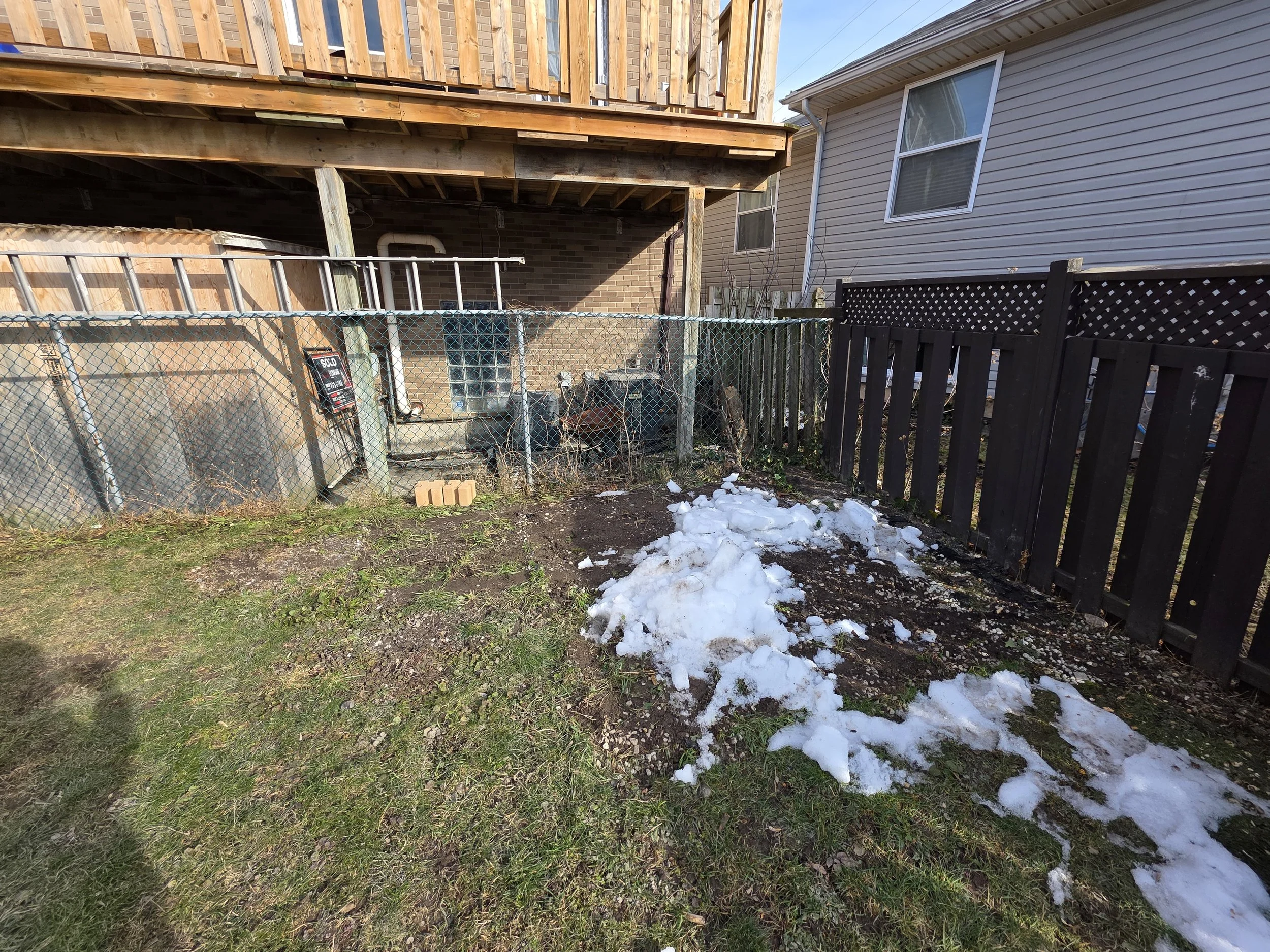 Backyard scene with snow patches on the grass, a wooden fence, a chain-link fence with a 'sold' sign, and a house with a beige exterior and white-framed windows.
