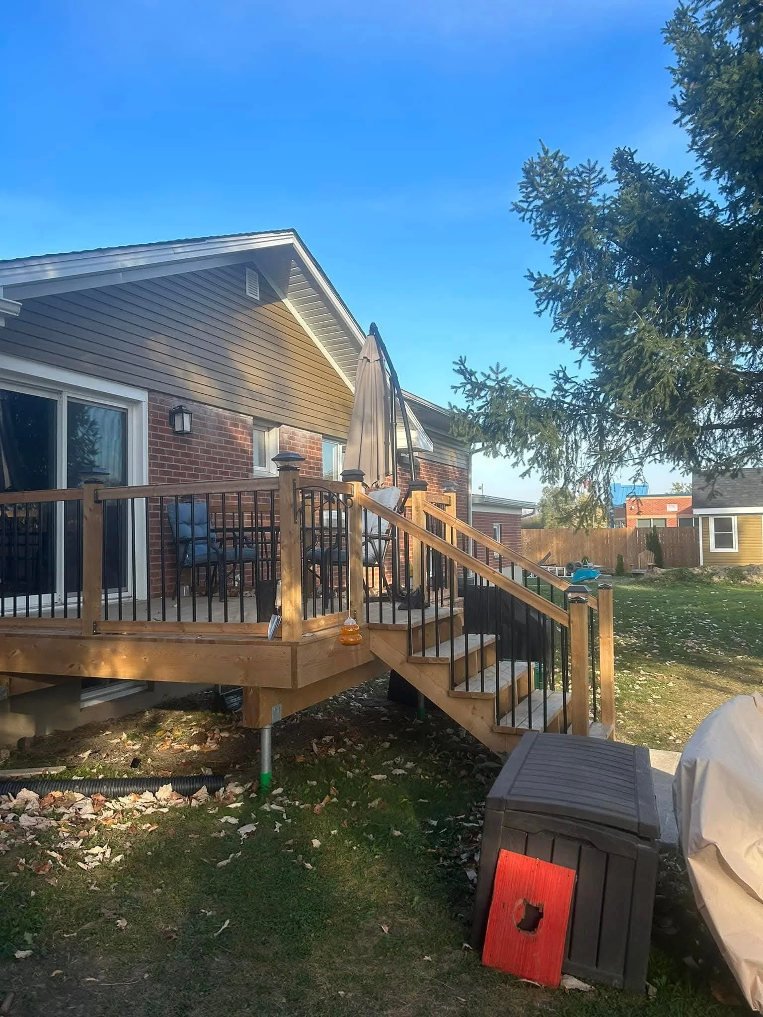 Backyard with a newly built wooden deck, patio furniture, a closed umbrella, trees, and a house with a brick and siding exterior under a clear blue sky.