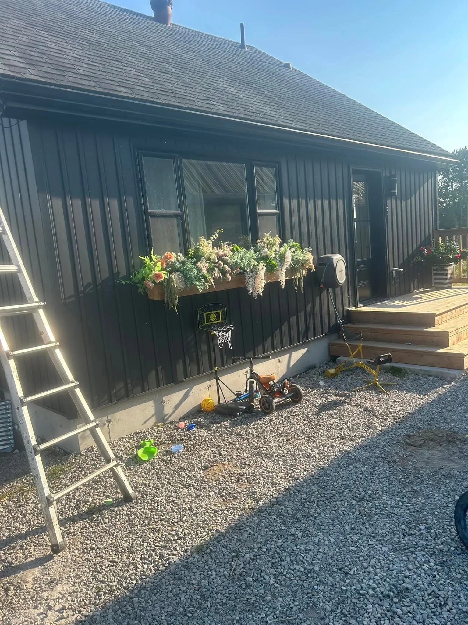 View of a house with black siding, front steps, and a window box with flowers. Children's toys and tools are scattered on a gravel yard in front of the house.