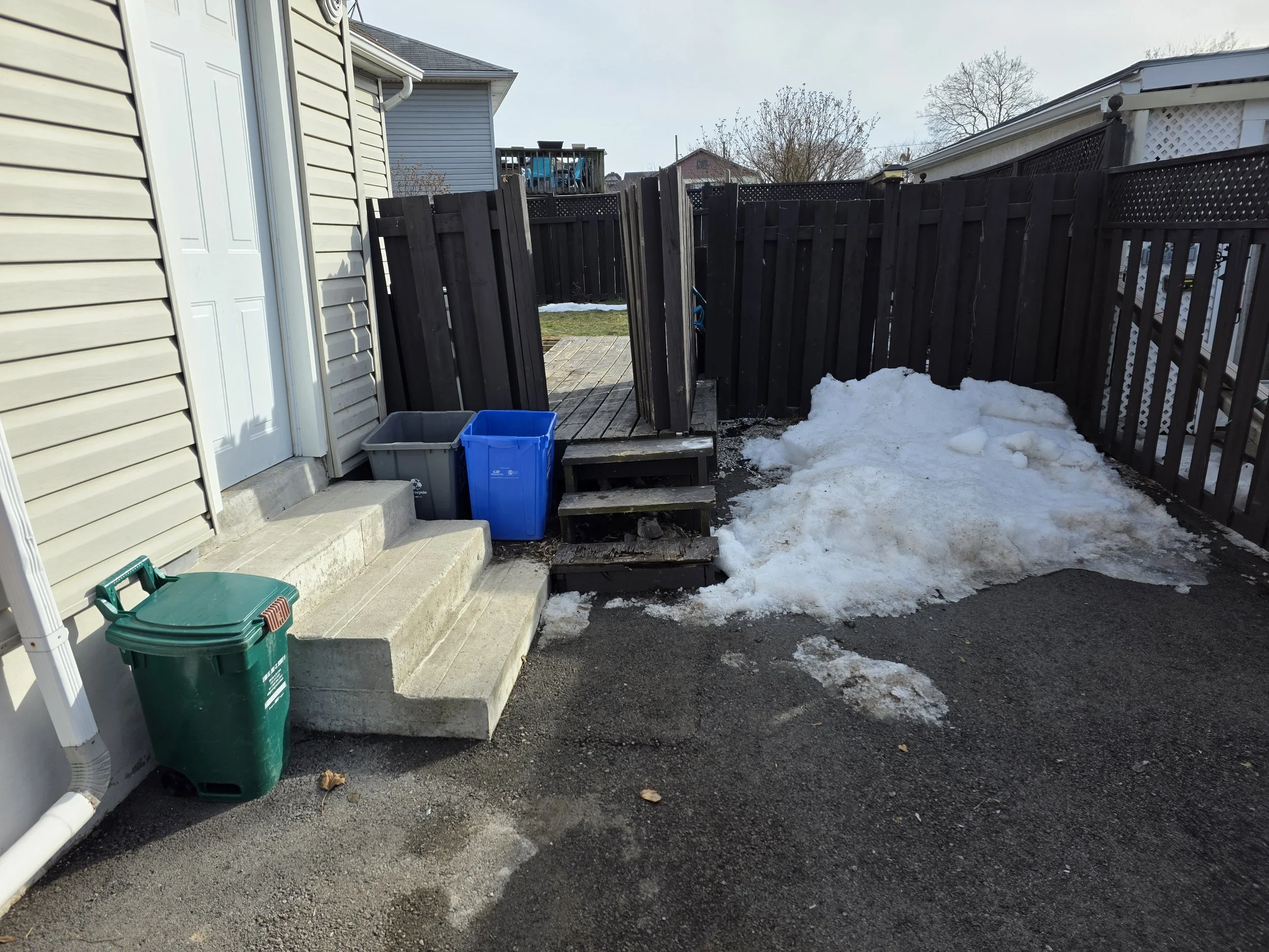 Backyard scene showing a wooden fence, steps leading to a wooden deck, snow on the ground, and trash bins near the steps.