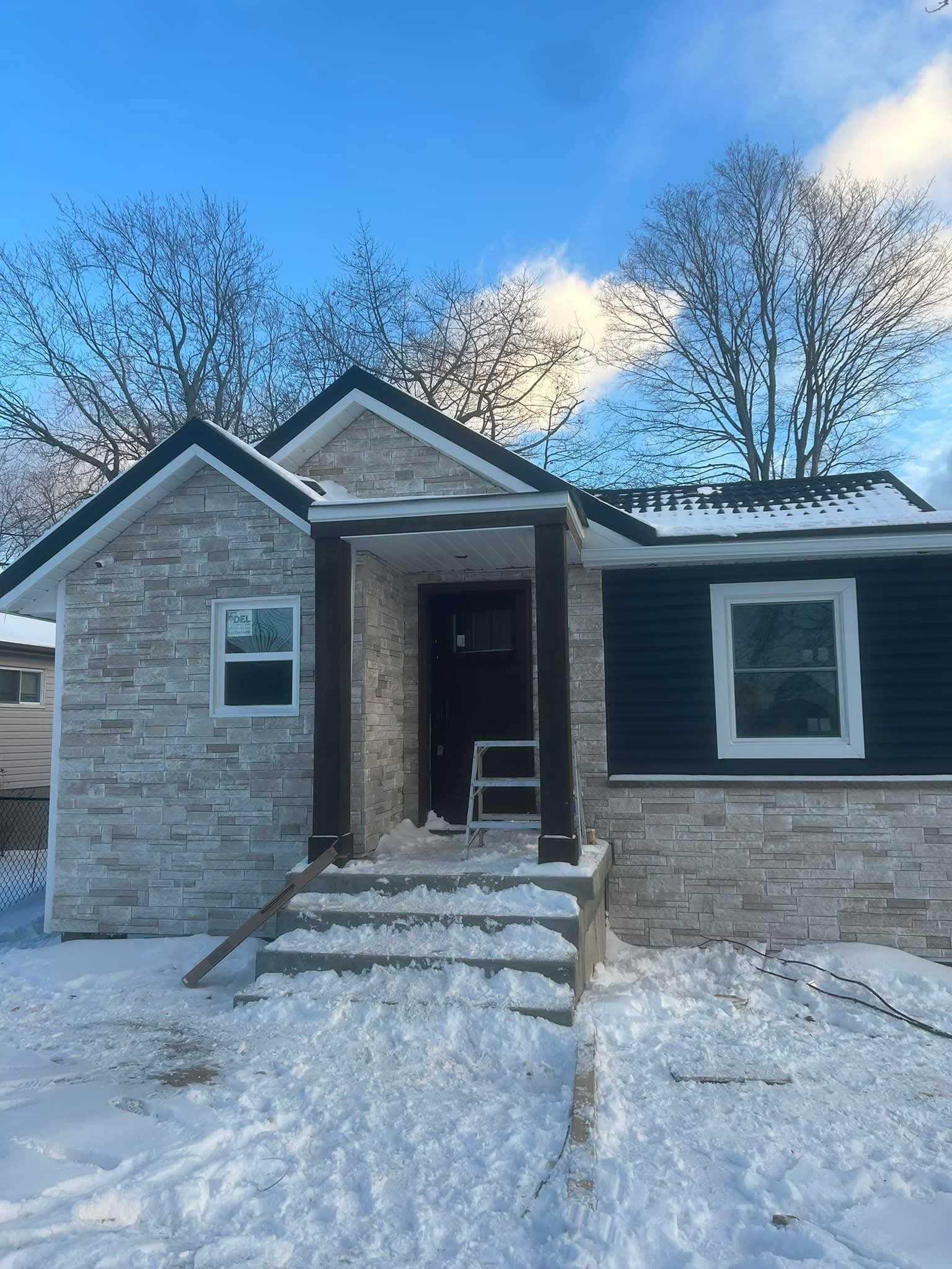 A house under construction with a front entrance stairway covered in snow, and a ladder leaning against it, with snow on the ground. The house has a stone exterior, a black roof with snow, and has two visible windows.