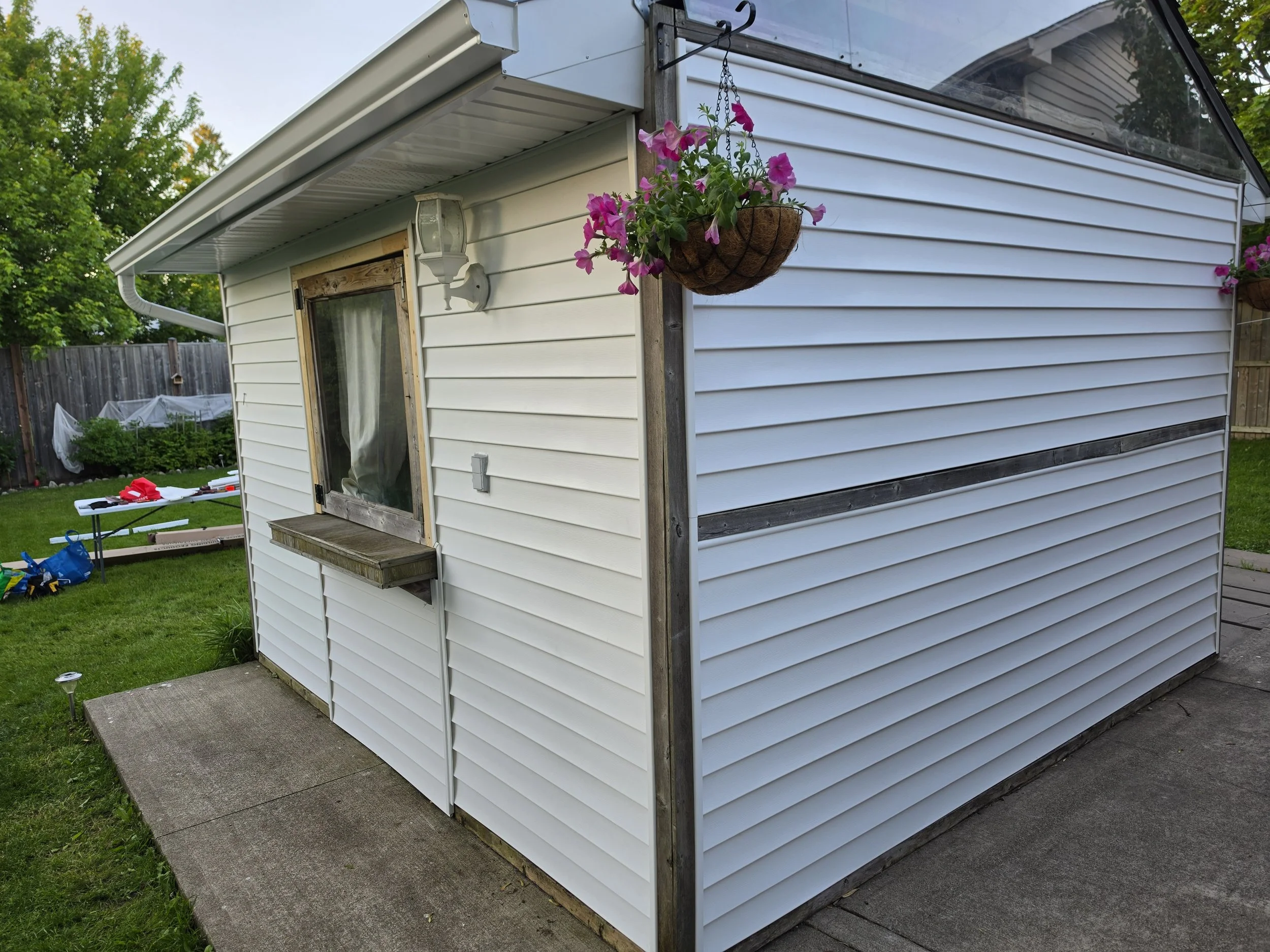 Exterior of a white house with horizontal siding, a sliding window, and a hanging basket of vibrant pink flowers, with a garden and wooden privacy fence in the yard.