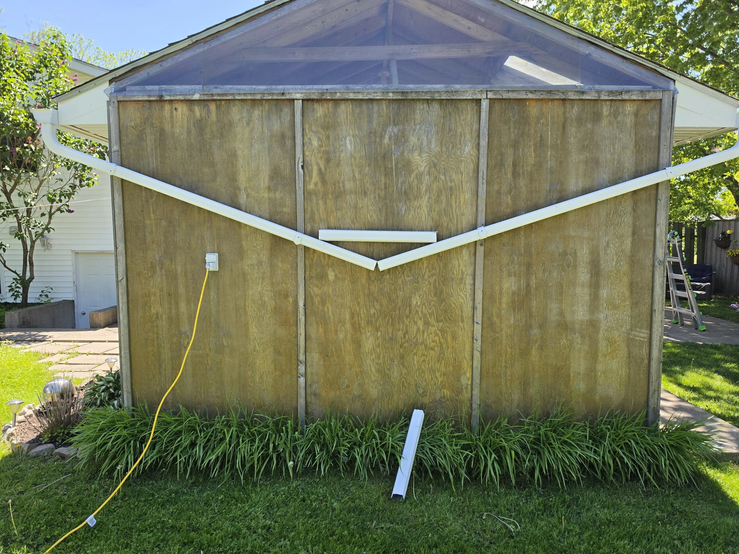 A backyard shed or small building under construction with unfinished wooden walls and white trim. There is a yellow extension cord on the ground and a white piece of trim leaning against the building. Green grass and plants are in the foreground, wit