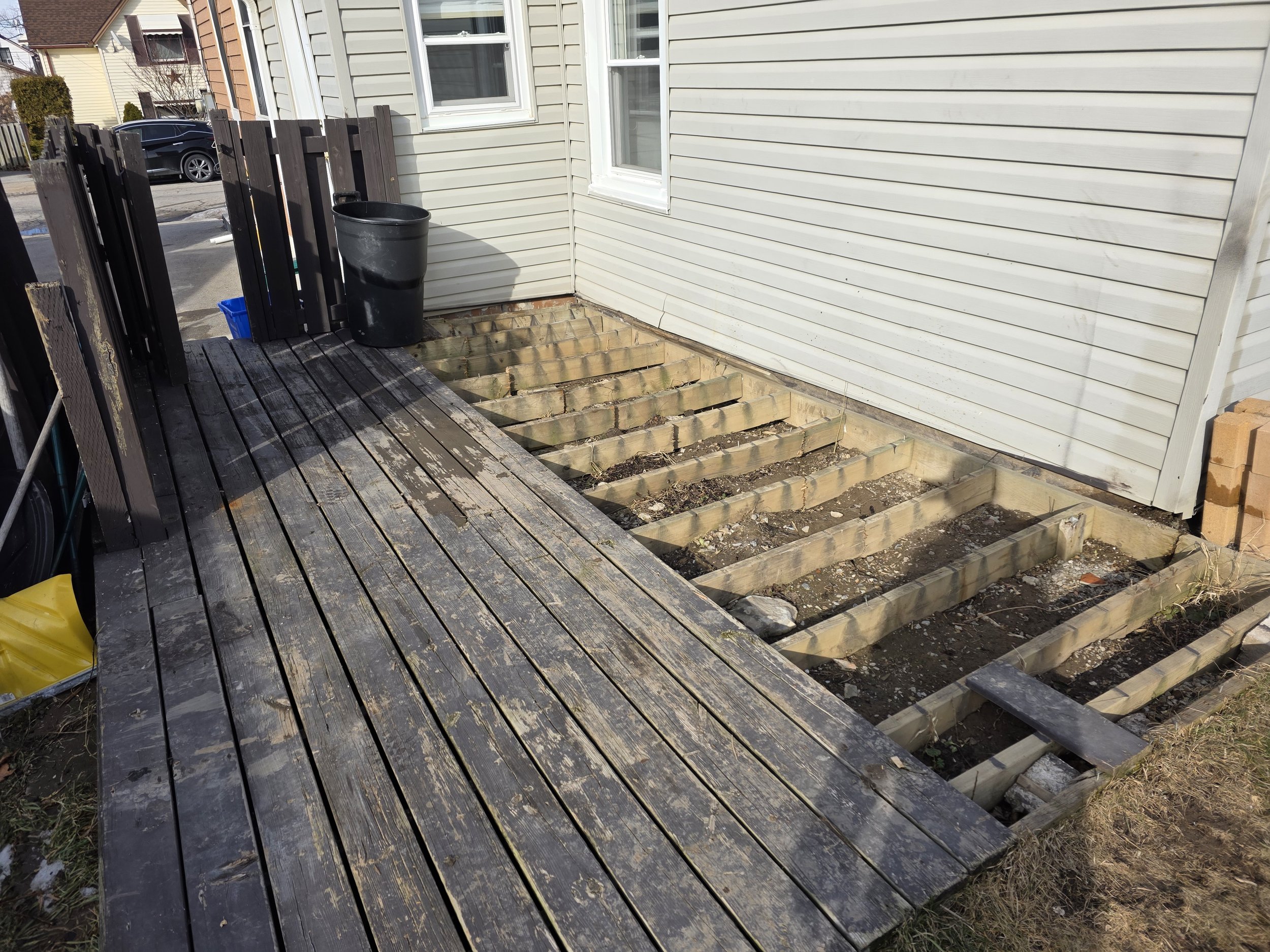 The image shows the exterior corner of a house with beige siding, two double-hung windows, and a section of a wooden deck that is under renovation, with missing floorboards and exposed joists. A black trash can and a blue recycling bin are placed on 