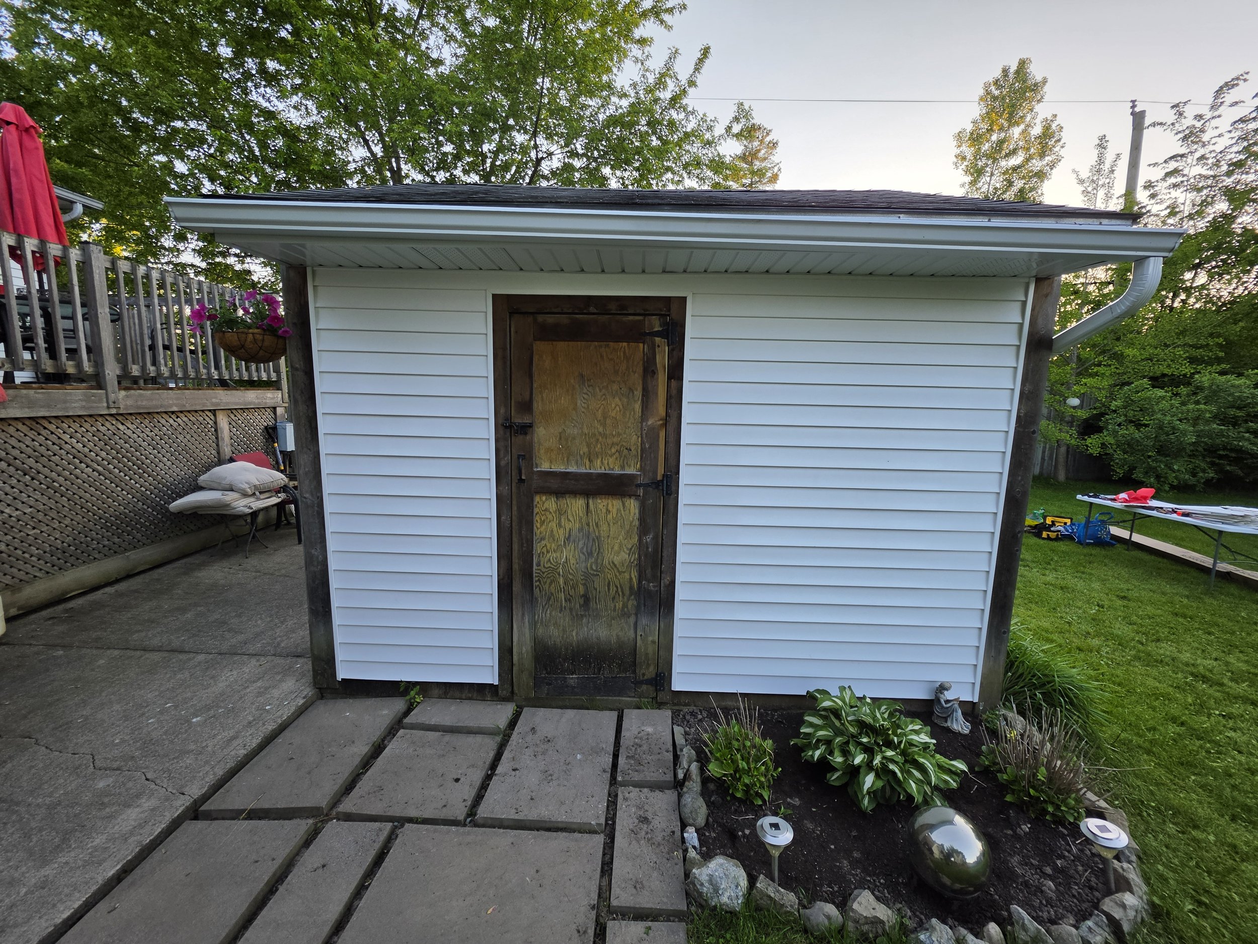 Backyard shed with white siding and a weathered wooden door, surrounded by garden and outdoor items, under a partly cloudy sky.