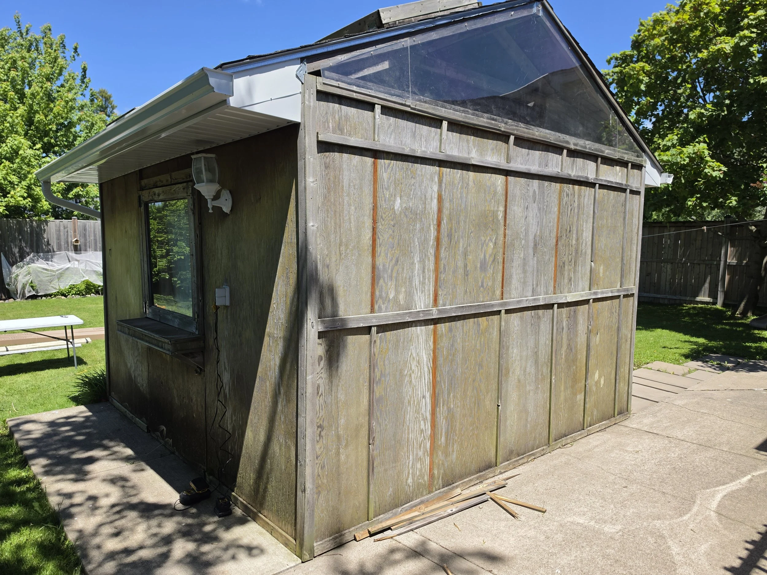 Backyard shed with weathered wooden planks, partially under repair with vertical and horizontal wood supports, next to a concrete patio and lush green lawn, with trees and a fence in the background under a clear blue sky.