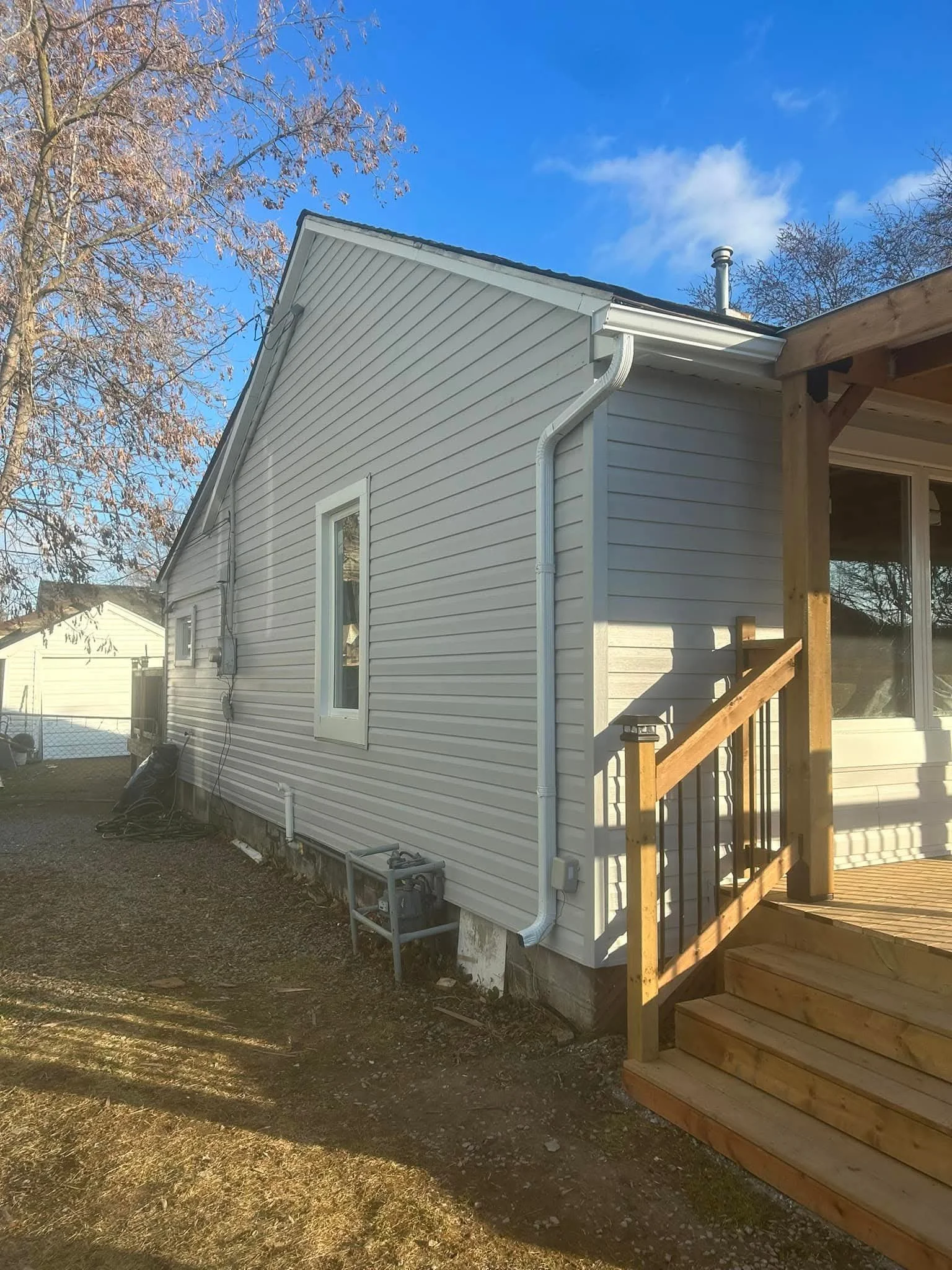 House with white vinyl siding, a small window, and a new wooden deck under construction. A tree with some remaining leaves is visible in the background under a clear blue sky.