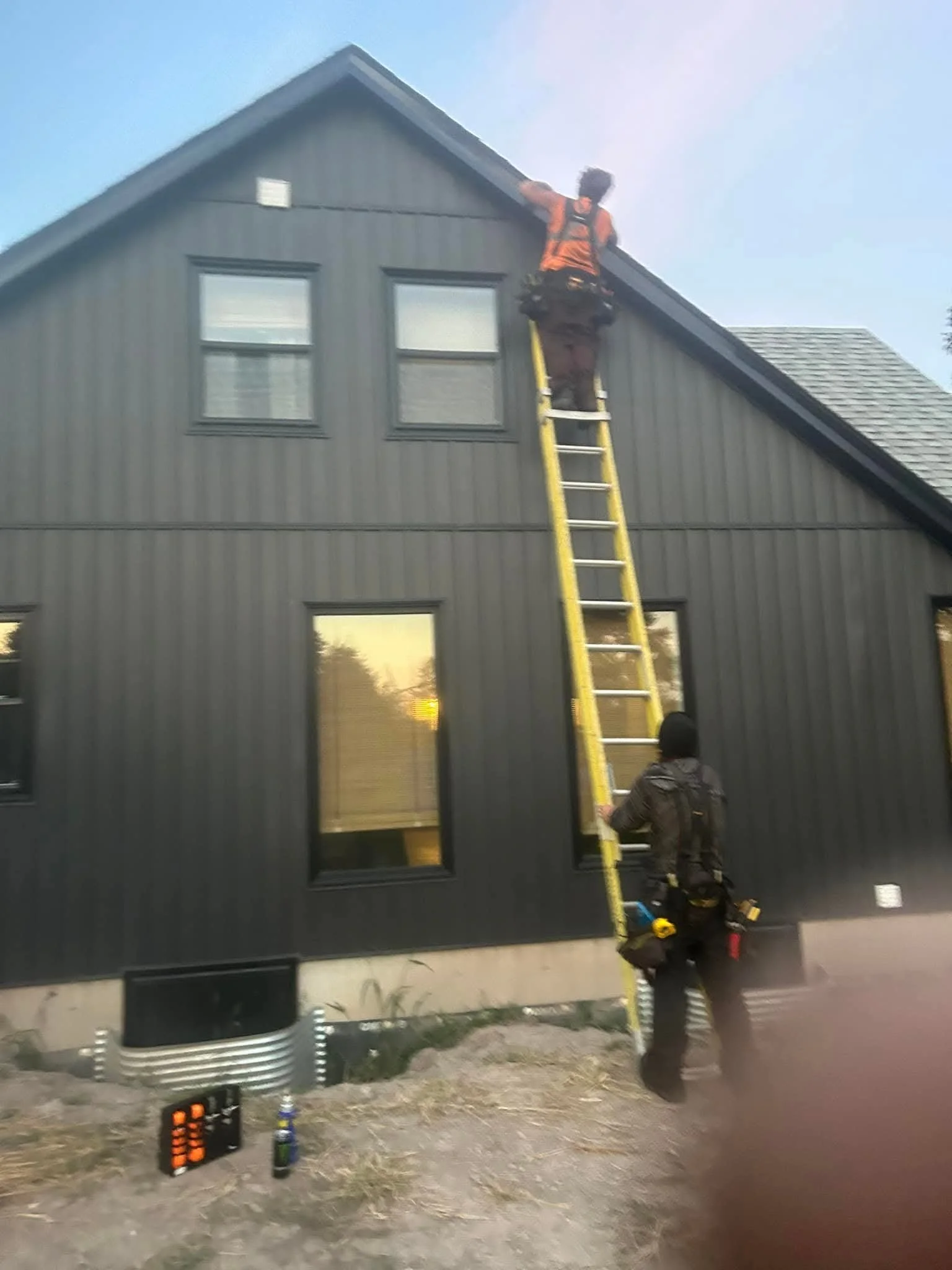 A person on a yellow ladder working on the roof of a dark gray house with two windows, while another person supports the ladder. Tools, a water bottle, and equipment are on the ground.