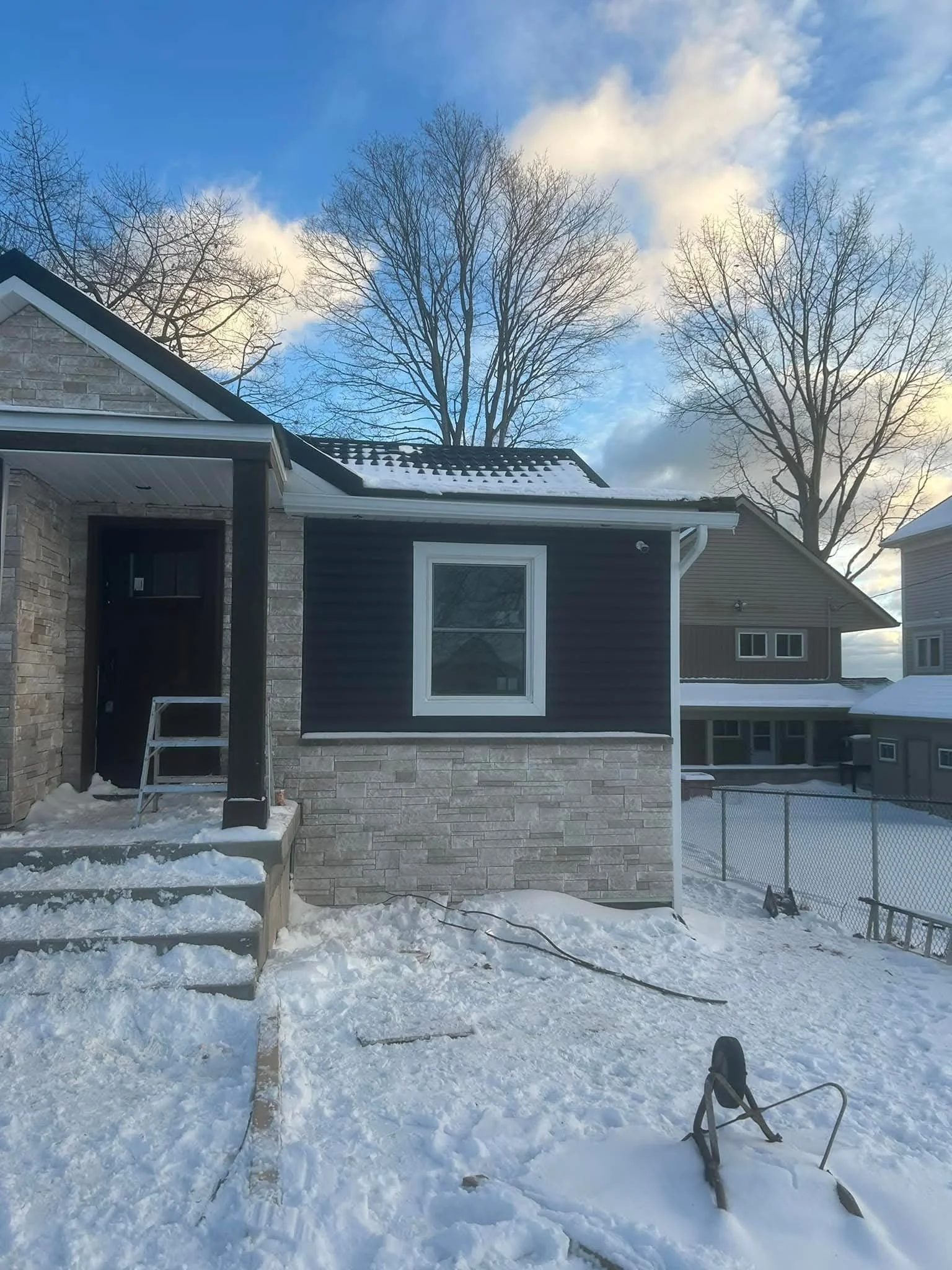 Backyard with snow-covered ground, house with brick and siding exterior, large window, and leafless trees against a cloudy sky.
