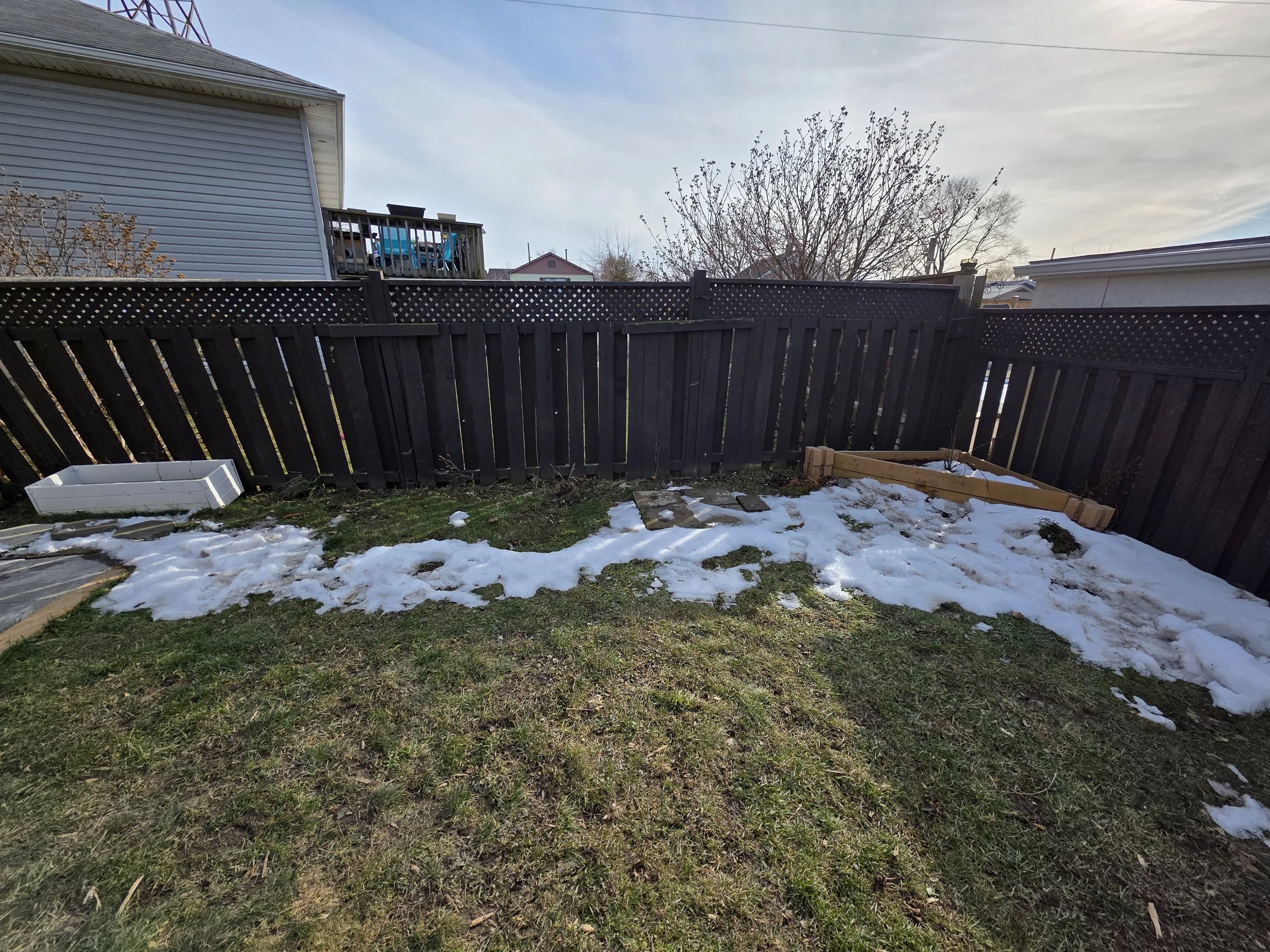 A backyard with a black wooden fence, patches of snow on the grass, a white planter box, and a wooden frame in the corner. Two houses and a leafless tree are visible in the background under a partly cloudy sky.