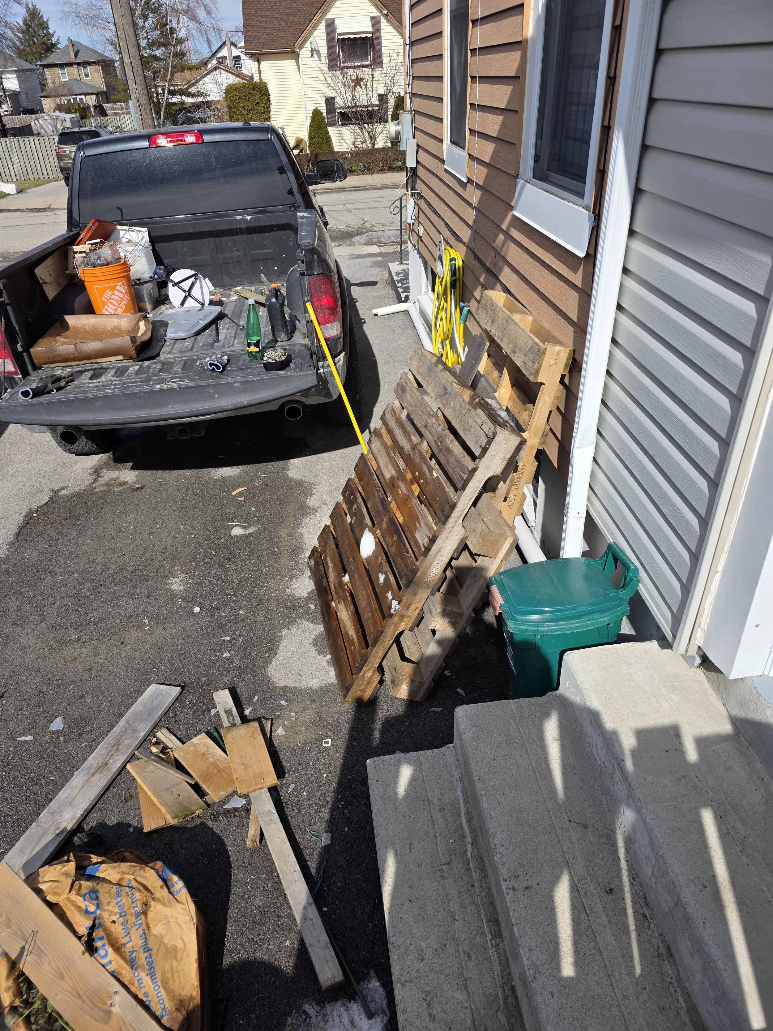 A black pickup truck parked outside a house, with various tools, a green bottle, and a Home Depot orange bucket in the truck bed. Next to the house, there are wooden pallets, yellow garden hoses, and a green storage container. The house has beige and