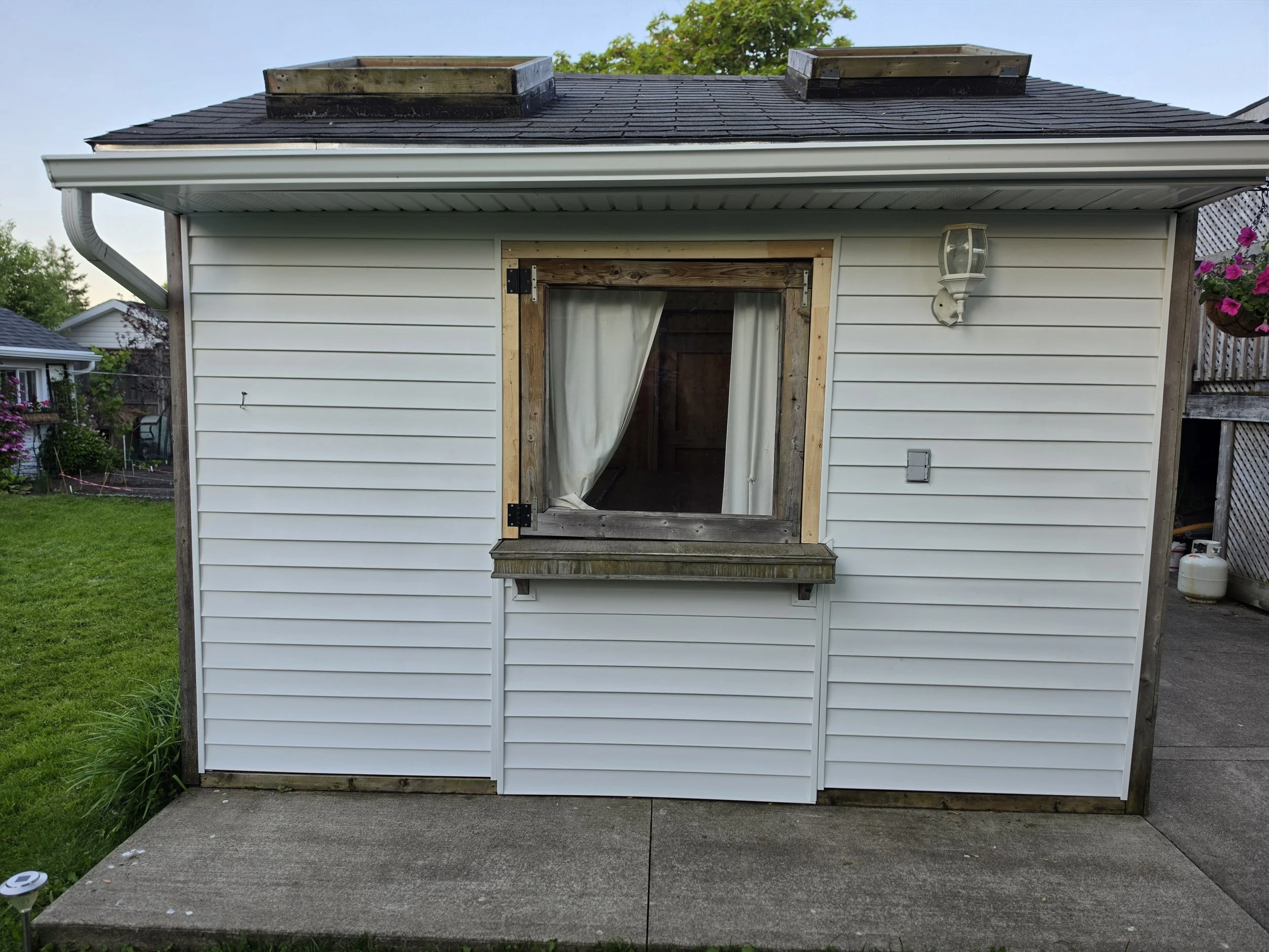 Back view of a small house or shed with white vinyl siding, a window frame without glass and a curtain, a wall-mounted outdoor light, and a concrete porch with a grassy yard nearby and neighboring houses in the background.