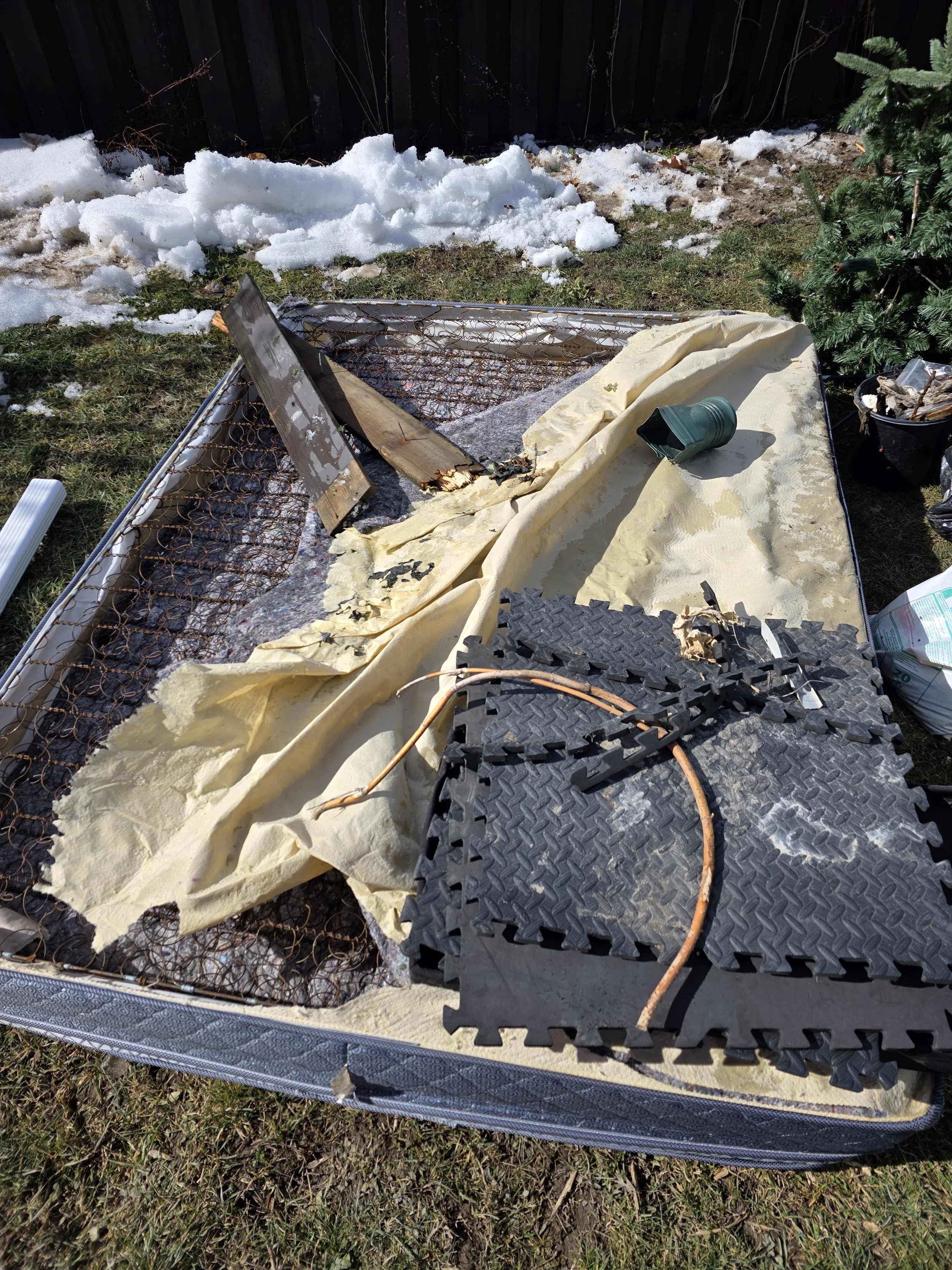 Disassembled metal bed frame with broken parts, yellow foam padding, a green plastic cup, and outdoor snow and grass, next to a Christmas tree outside.