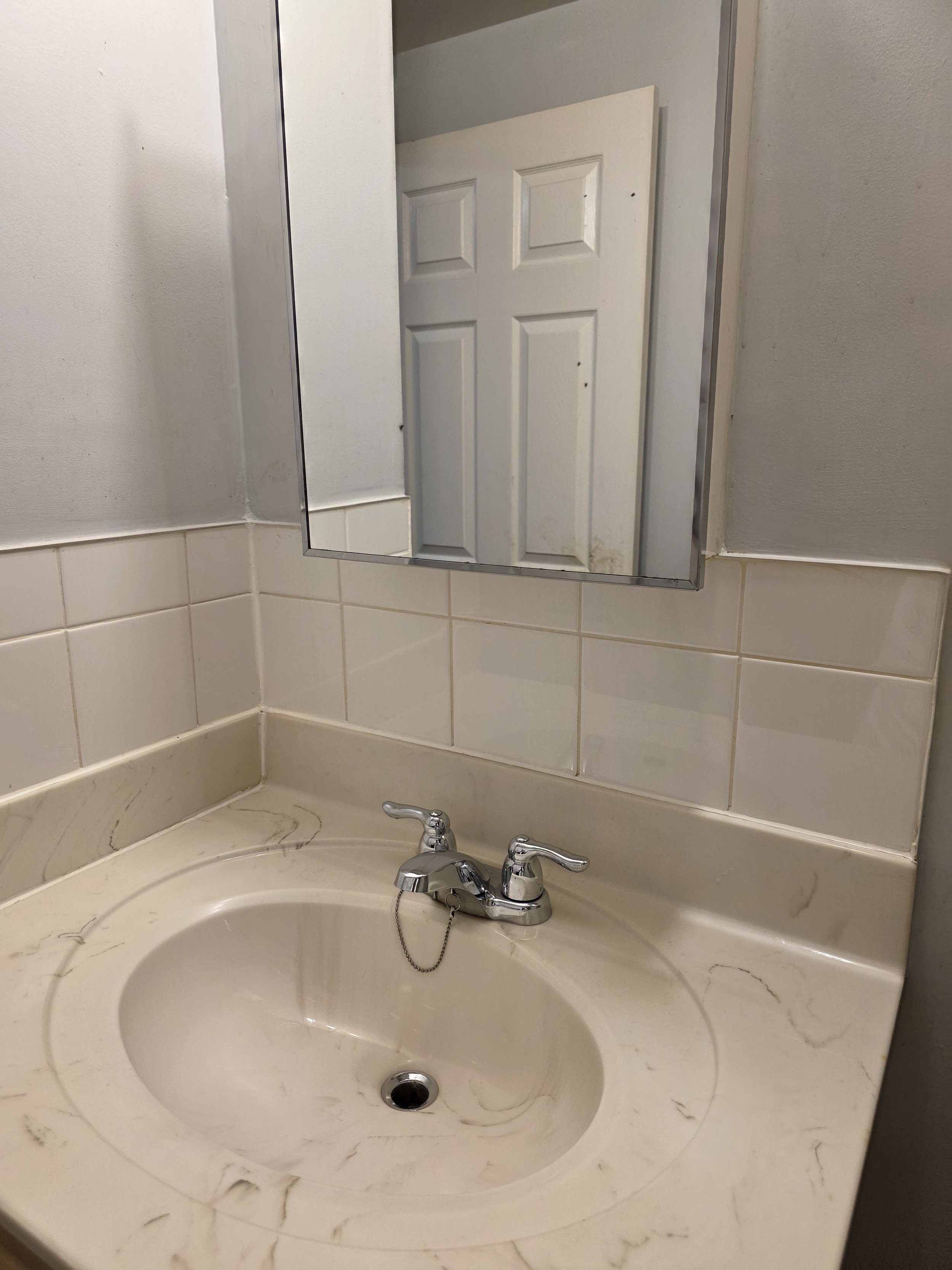 A bathroom sink with a marble-patterned countertop, a chrome faucet, and a mirror reflecting a closed white door with six panels.