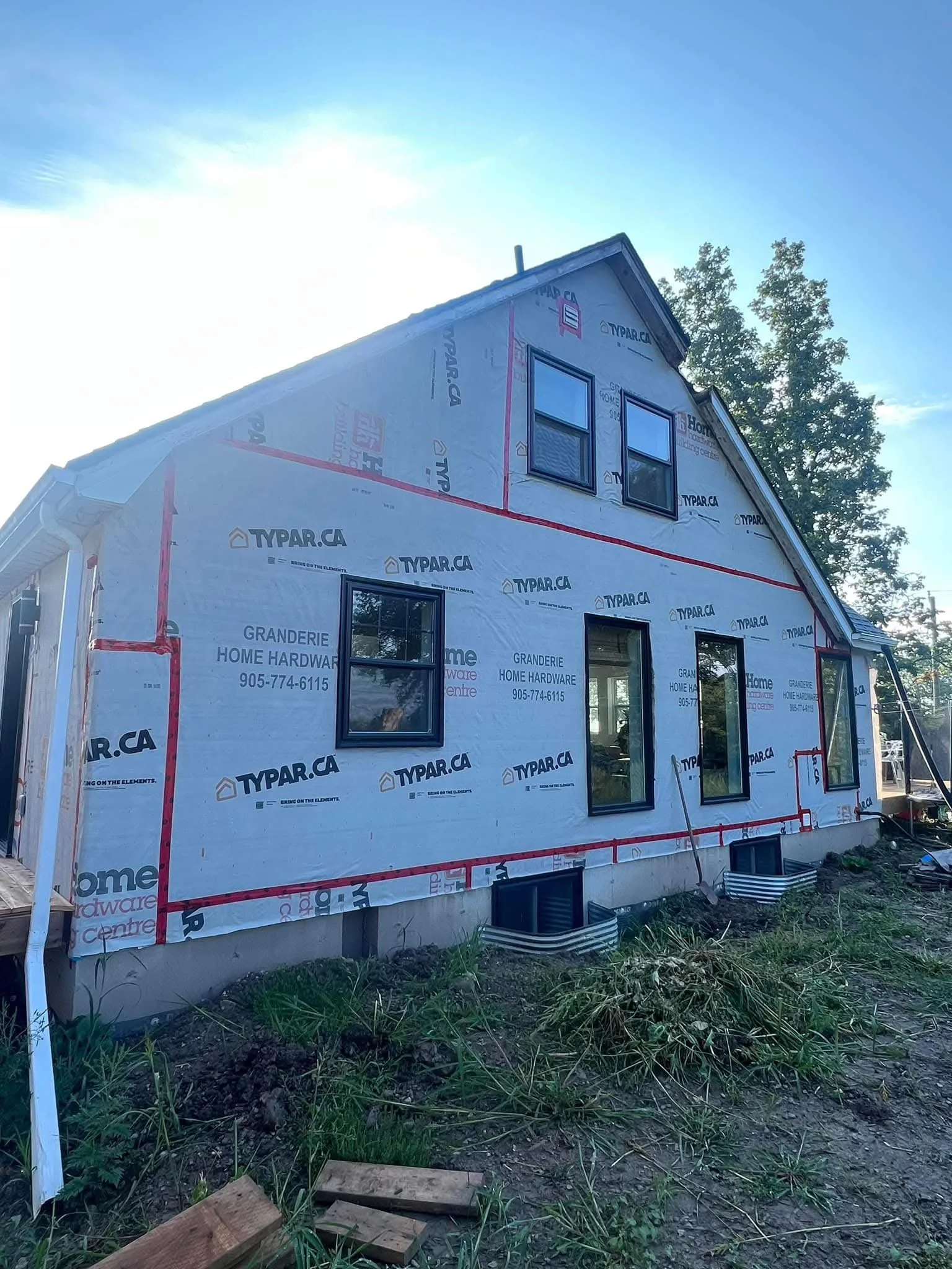 A house under construction with unfinished exterior walls covered in weather barrier wrap. Several windows are installed, and the roof is partially completed. Construction materials and tools are visible on the ground outside.
