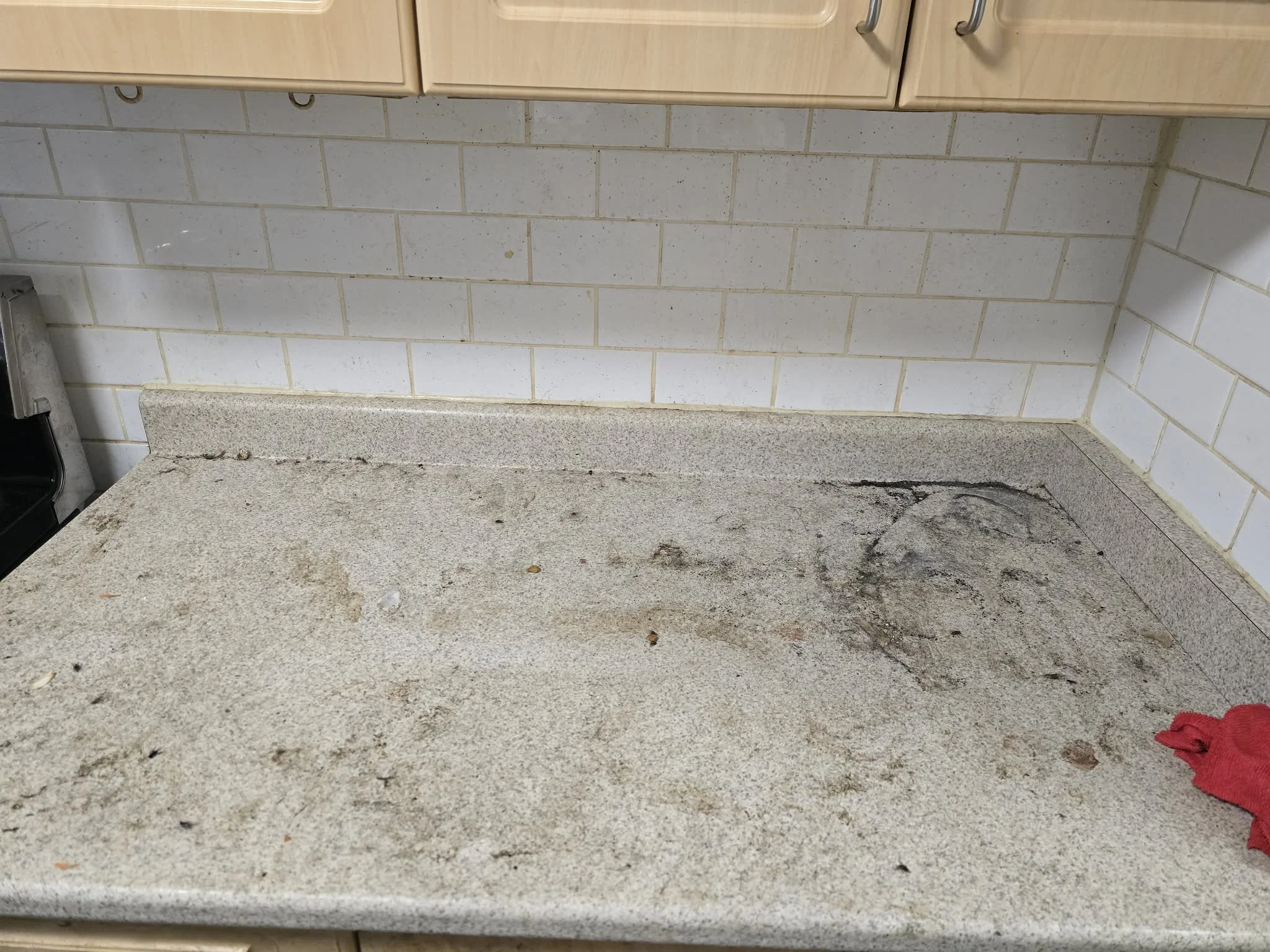 Close-up of a kitchen counter with a granite surface and a backsplash of off-white subway tiles. There is a red cloth on the lower right corner and some dirt or debris on the counter.