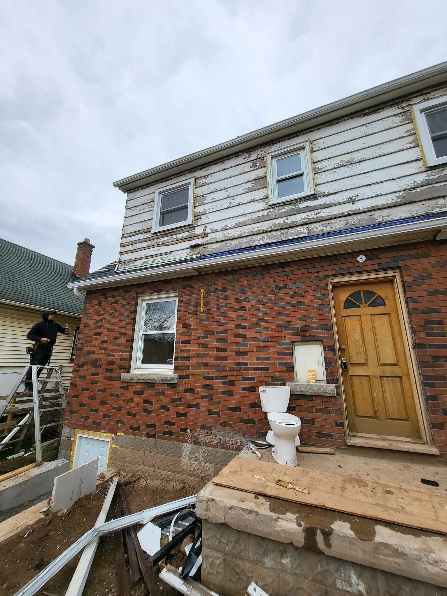 A two-story house under renovation with a brick lower level and a worn white wooden upper level. The house has three windows on the upper floor and one on the lower. Construction debris, tools, and a ladder are visible in the yard, and an outdoor toi