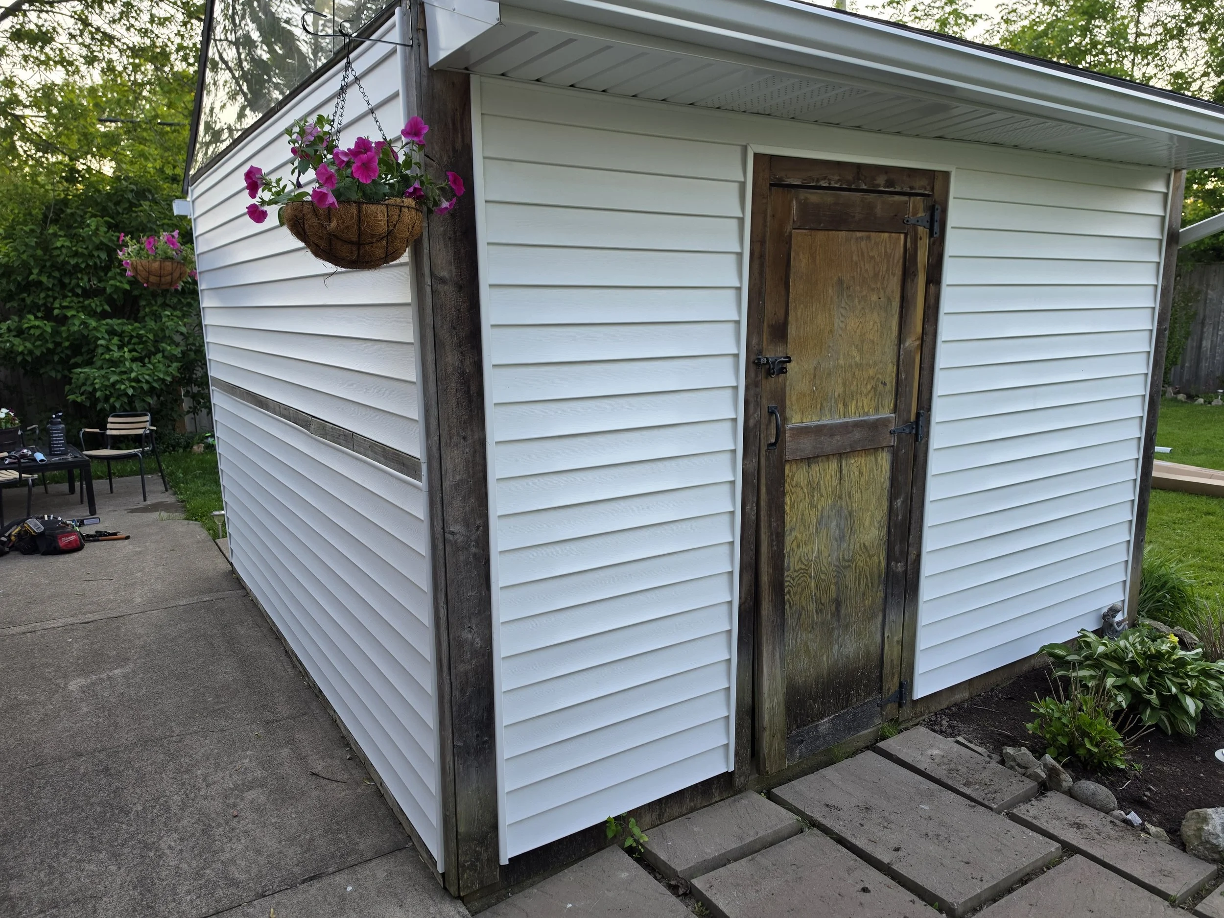 A small shed with white vinyl siding, wooden framing, and a wooden door secured with a latch hinge. Two hanging flower baskets with pink flowers are attached to the shed, with a landscaped backyard and patio furniture in the background.