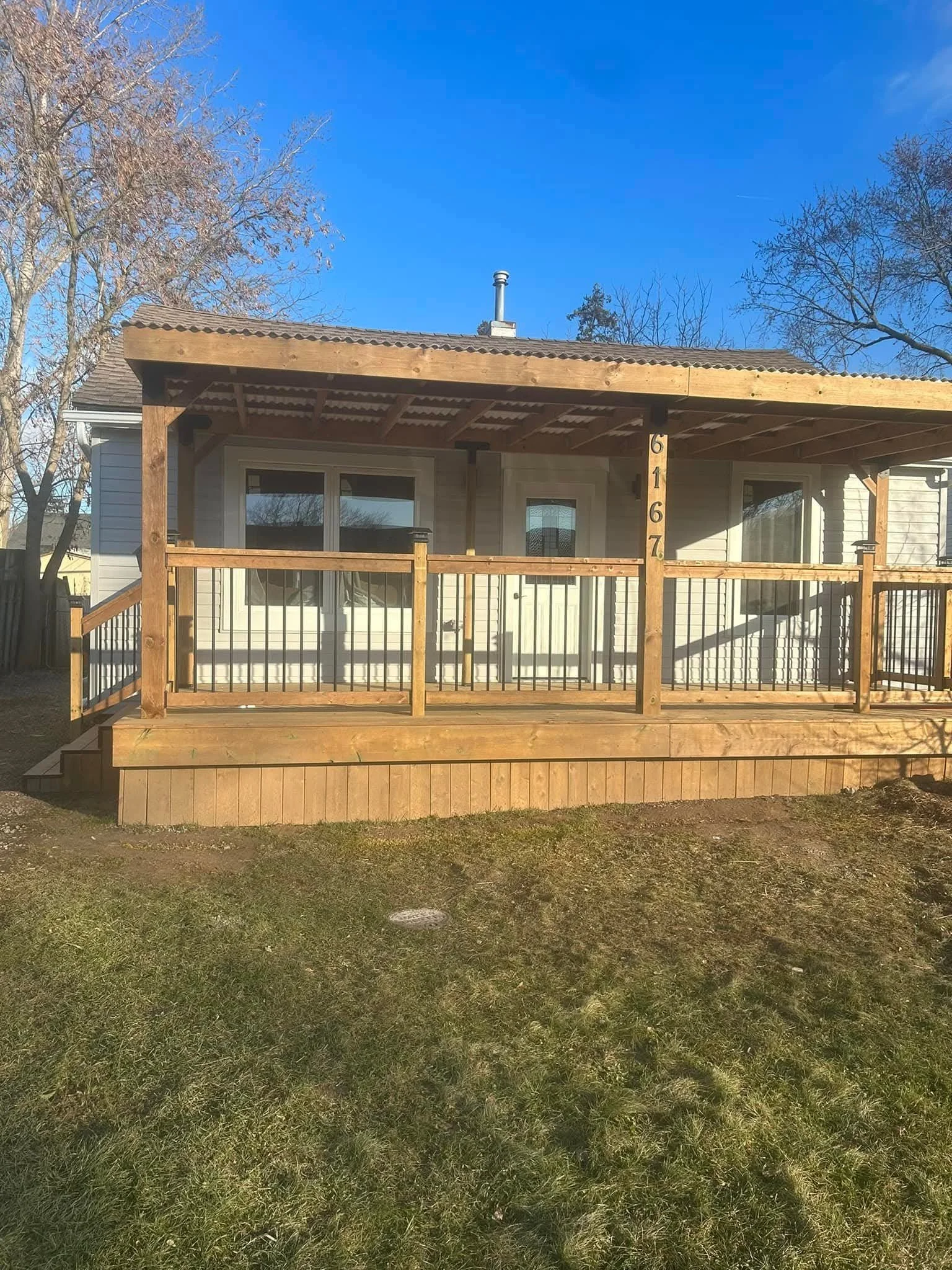 A house with a wooden porch and a white exterior, surrounded by trees, with a clear blue sky overhead.