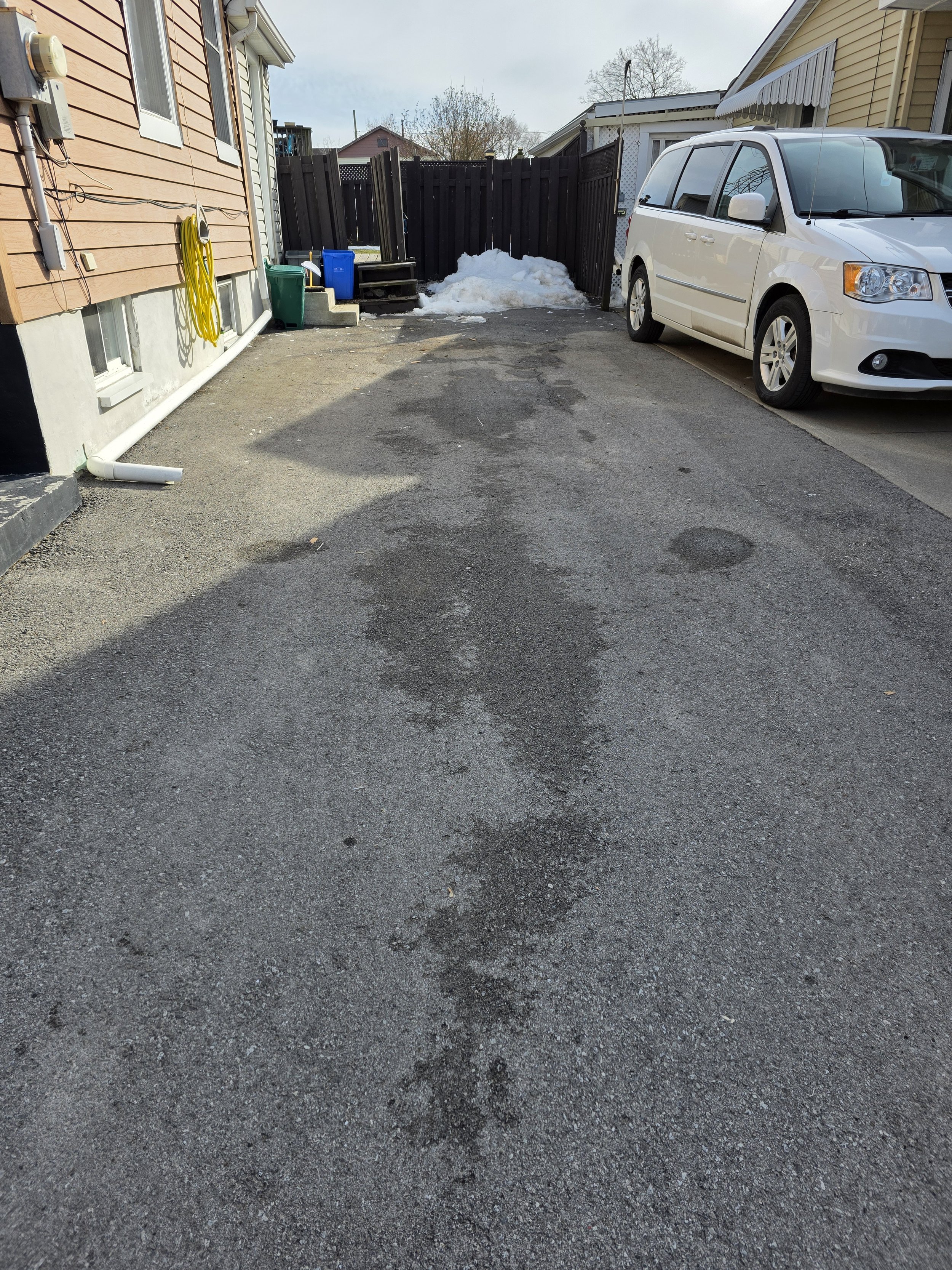 Driveway with patchy asphalt, some snow piles at the end, and a white minivan parked on the right side. Residential houses with siding are visible in the background.
