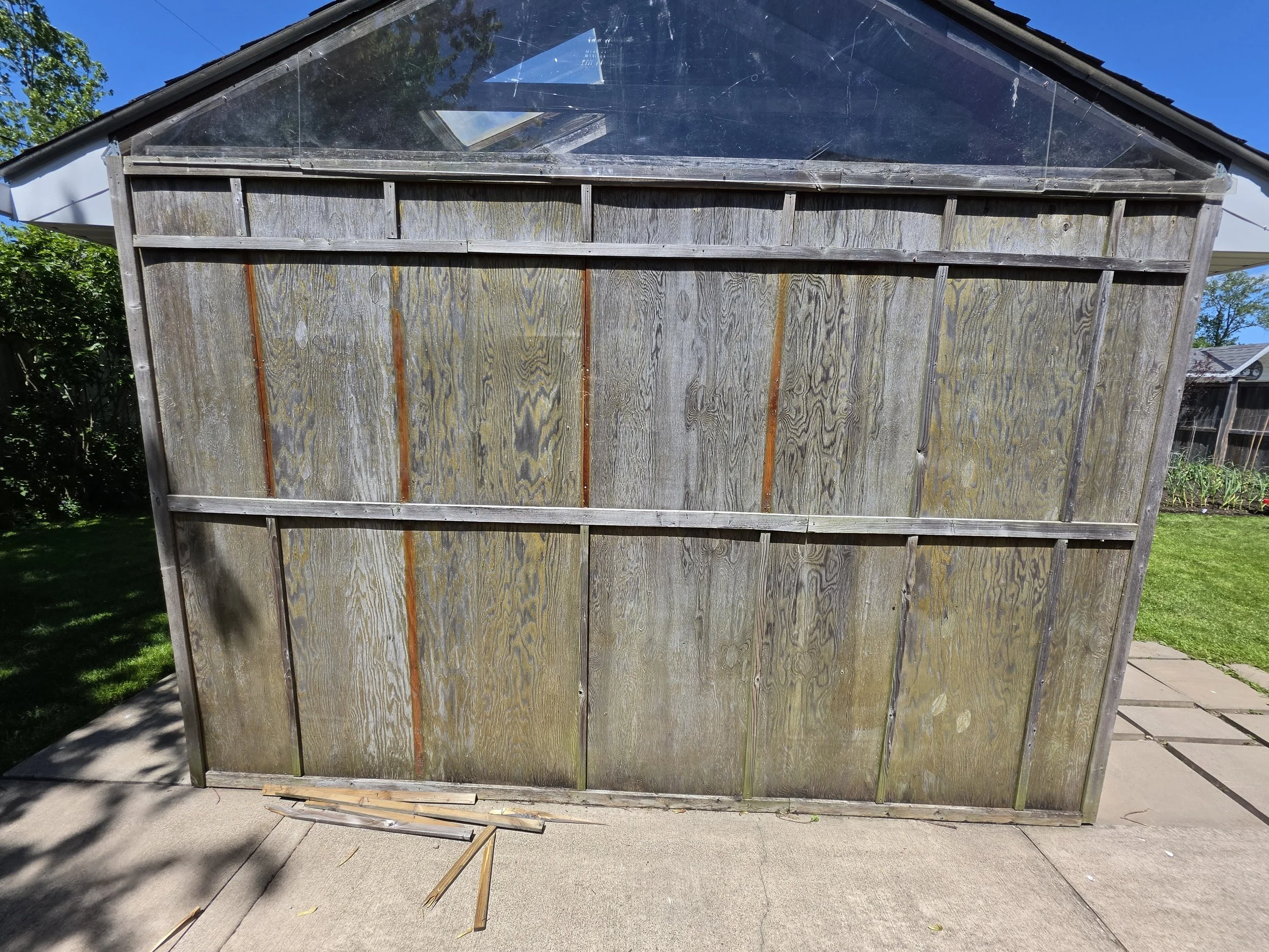 A large wooden structure, possibly a discarded shed or fence, with weathered vertical planks, some of which are damaged or missing. It is located outside on a concrete surface, with trees and a clear blue sky in the background.