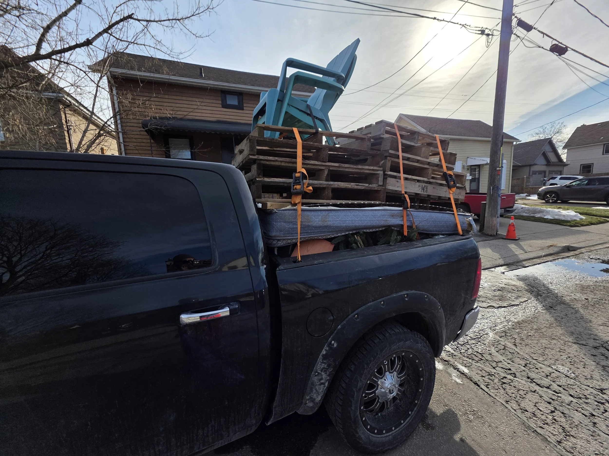 The image shows the back of a black pickup truck with its bed loaded with wooden pallets and a couple of plastic chairs. The pallets and chairs are secured with orange straps. There is a street scene with houses, a utility pole, and cars in the backg