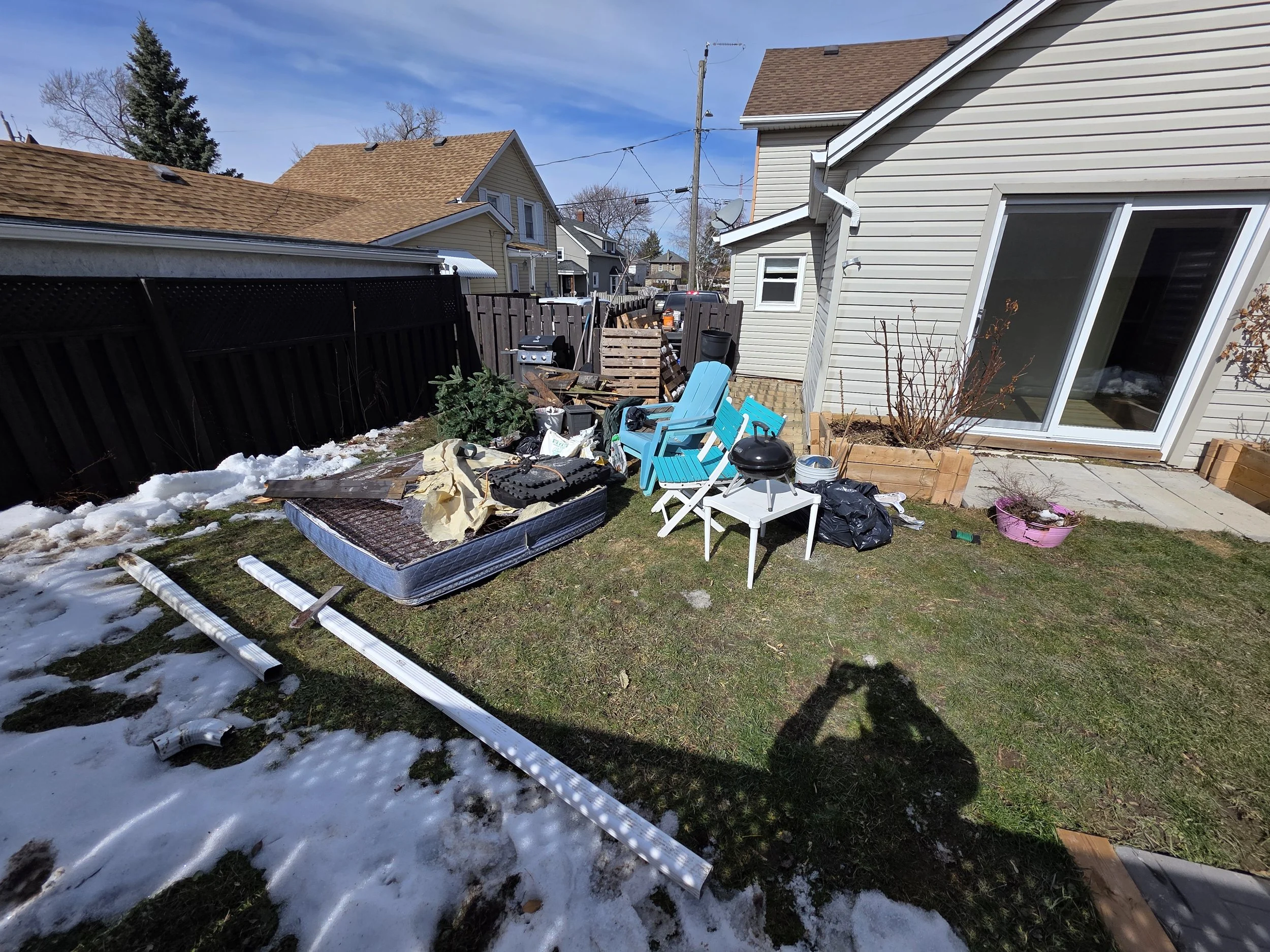 Backyard scene with partially snow-covered grass, scattered debris including a disassembled white ladder, blue and white chairs, a pink planter, a barbecue grill, and a pile of wooden pallets. Surrounding houses with brown and beige siding, fenced ya