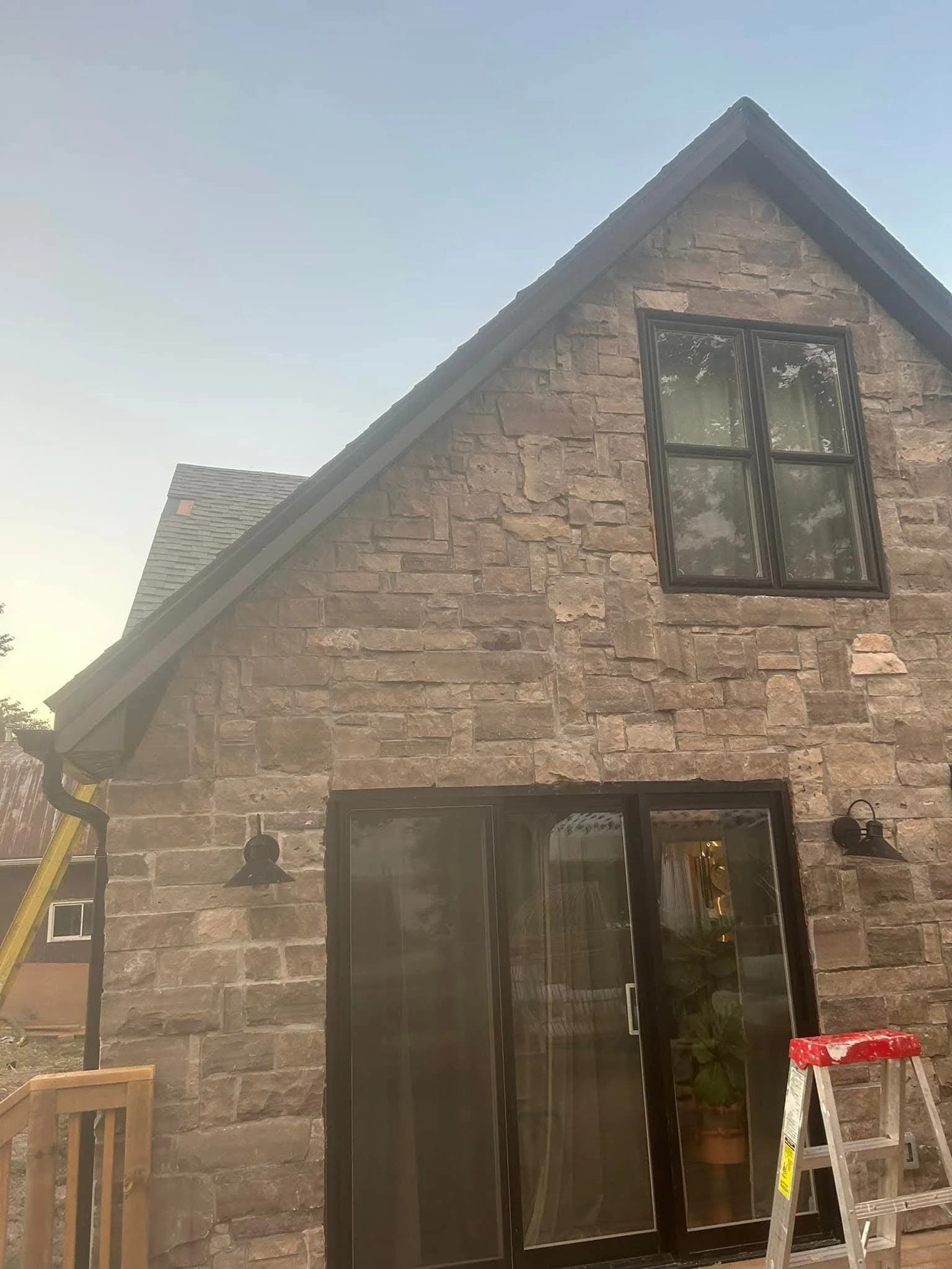 A house with a stone exterior and a gabled roof under construction, featuring large black-framed windows and outdoor wall-mounted lanterns, with a ladder and construction materials visible in the yard.