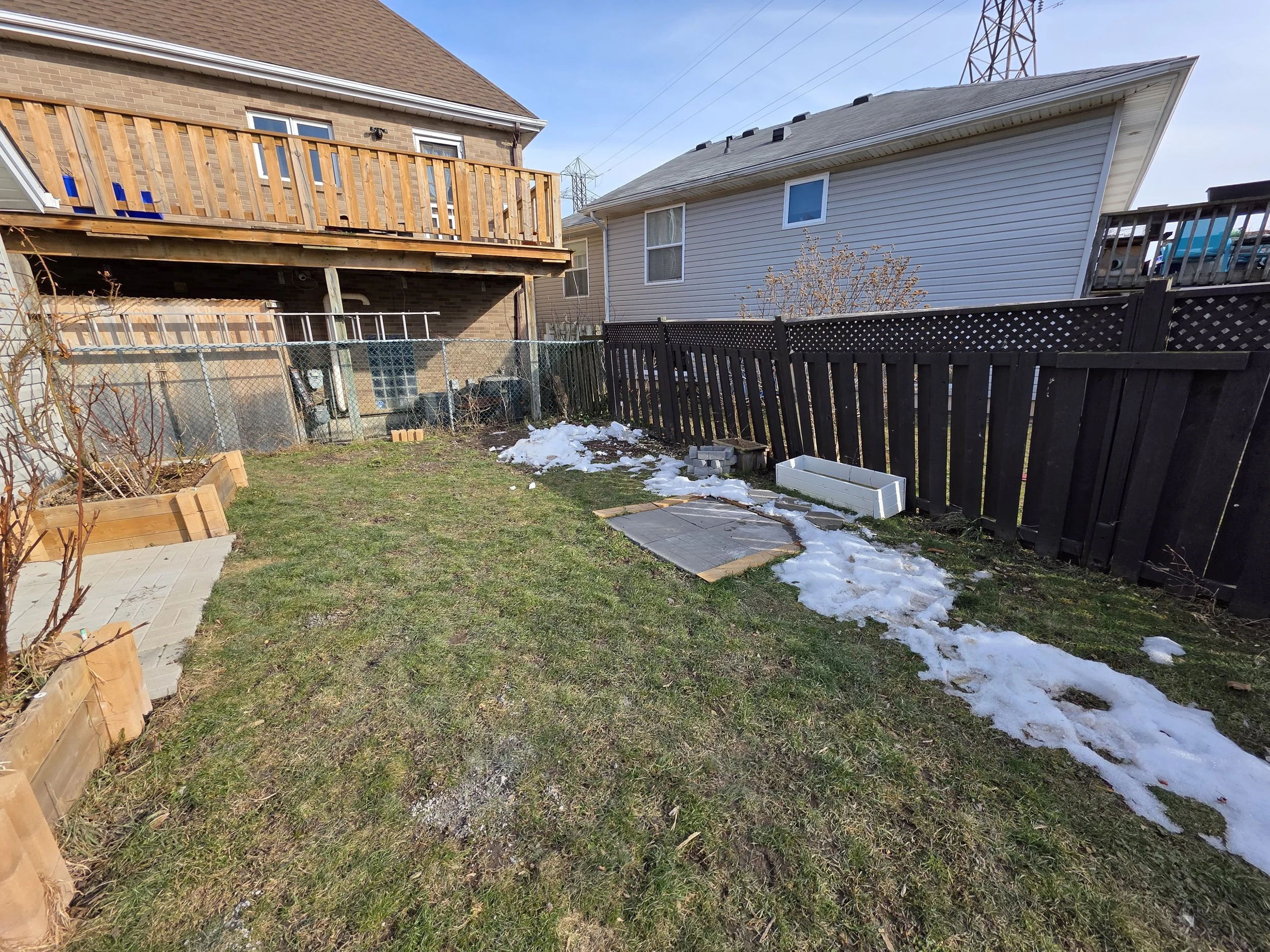 Backyard scene with a wooden deck under construction, a fence, a small patch of snow on the ground, and neighboring houses with a power line tower in the background.