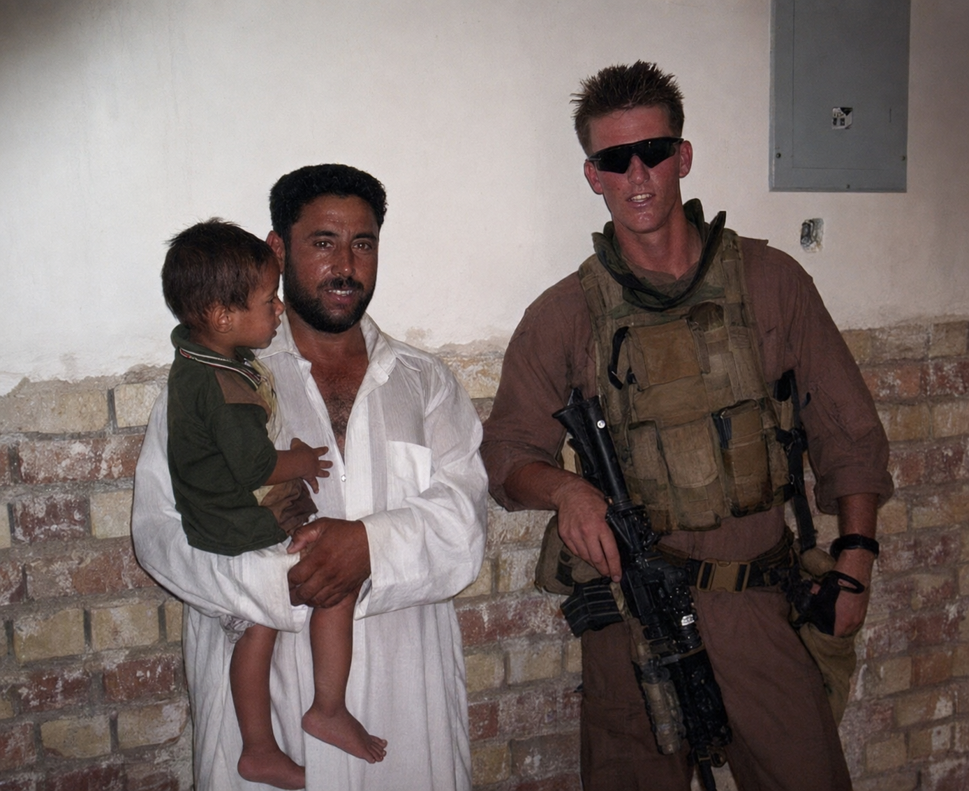 A man holding a child standing next to a soldier in military gear, against a brick wall background.