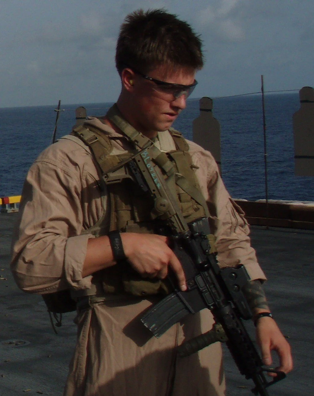 A soldier dressed in tan military uniform, holding an automatic rifle, standing on a ship with the ocean and sky in the background.