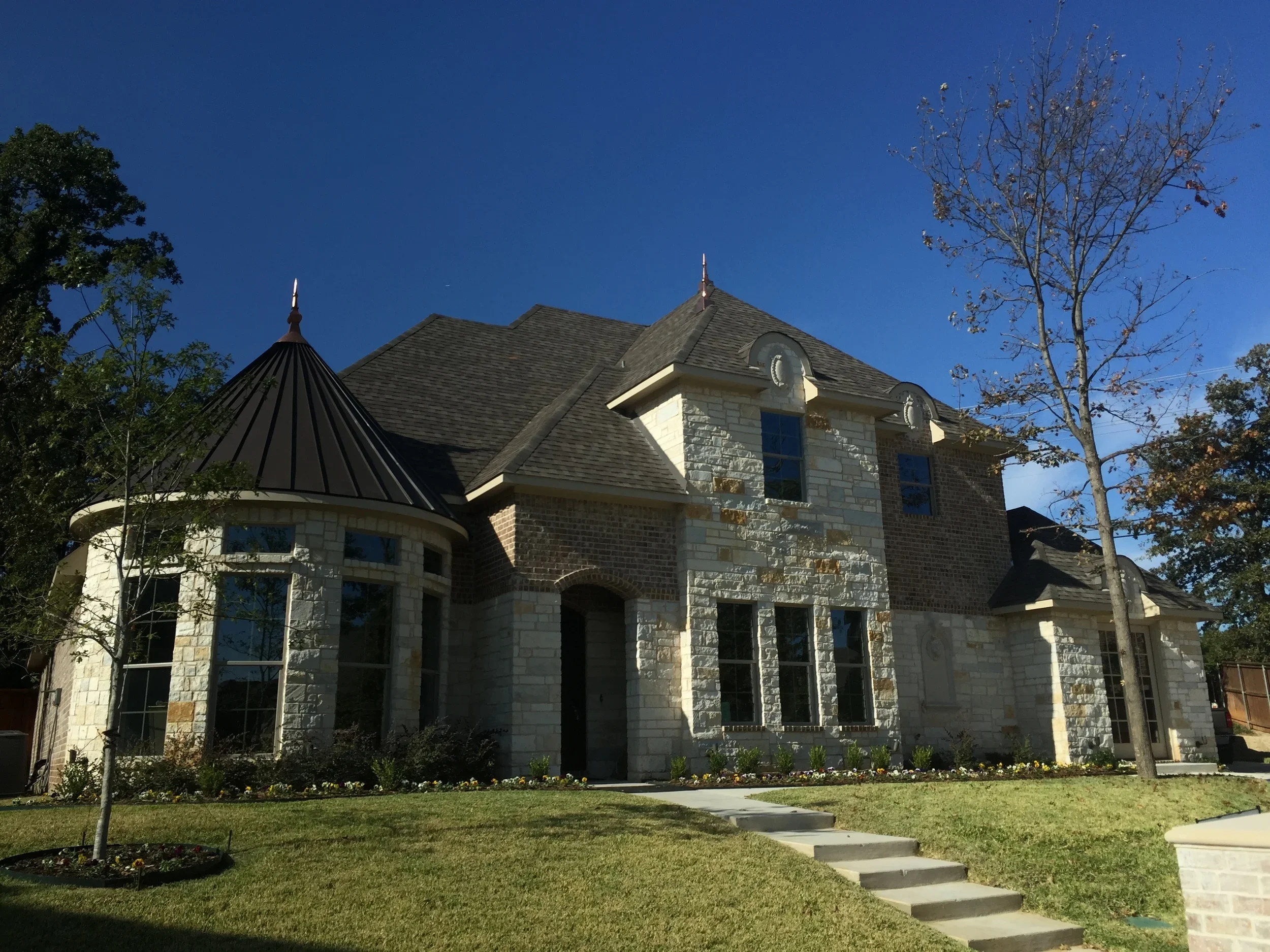 Front view of a large house with brick and stone exterior, multiple windows, a pointed roof, and a small front yard with a sidewalk, steps, and trees, one of which has sparse leaves, against a clear blue sky.