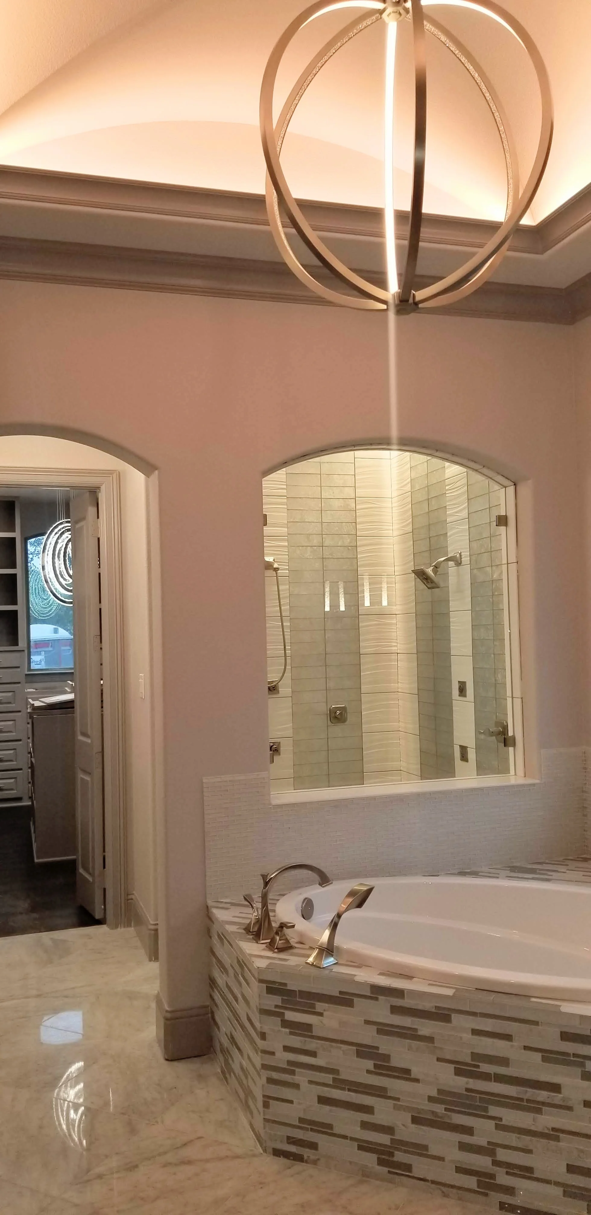 Spa style master bath in Carrollton complete with a built-in tub, custom tile work, a modern chandelier as well as an arched glass shower viewing wall.