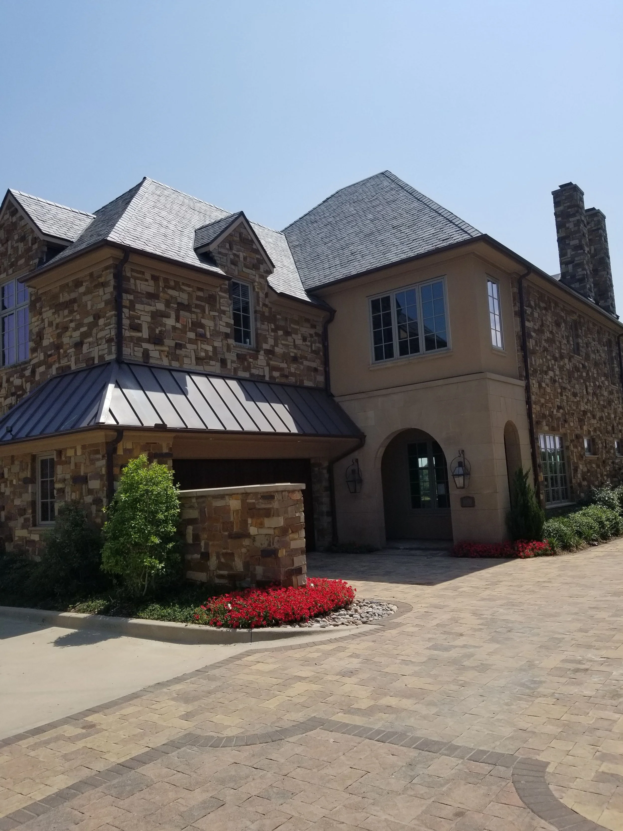 Stunning exterior of a custom luxury home in McKinney inspired by English Country style, with a blend of multi-colored stone, stucco, as well as metal roofing and custom landscaping adorning the driveway.