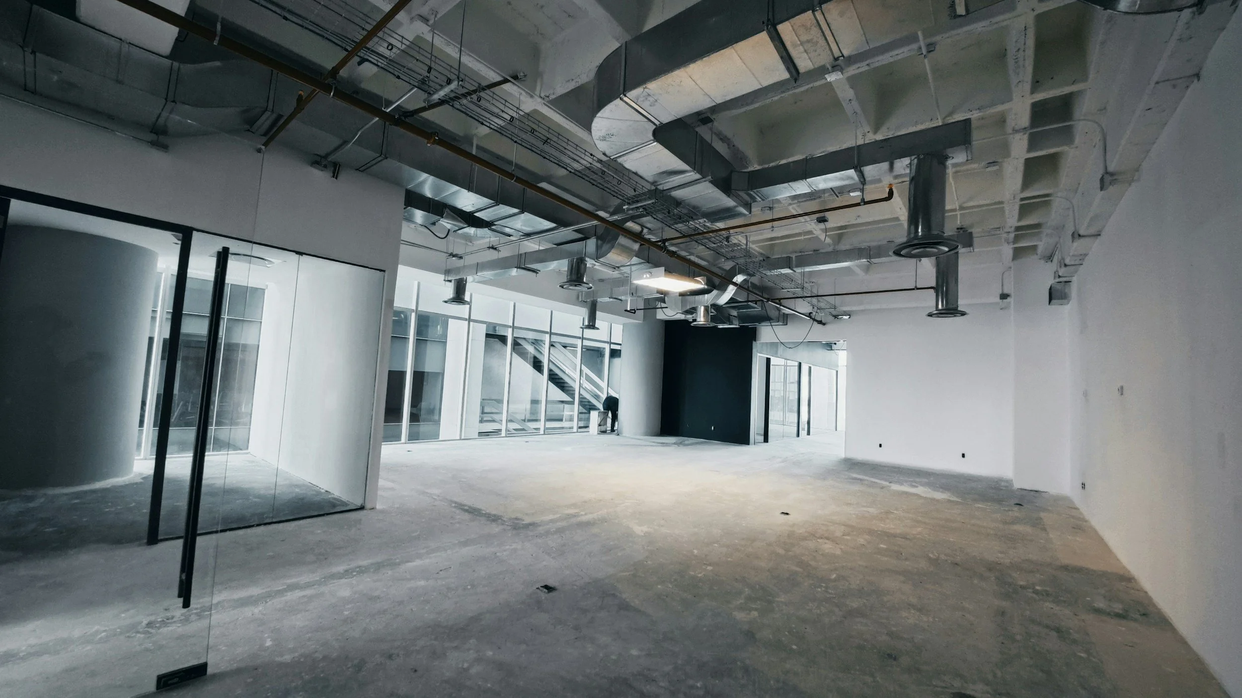 An unfinished interior space under construction with exposed ceiling pipes, ducts, and wiring, large windows, and a partially completed floor.