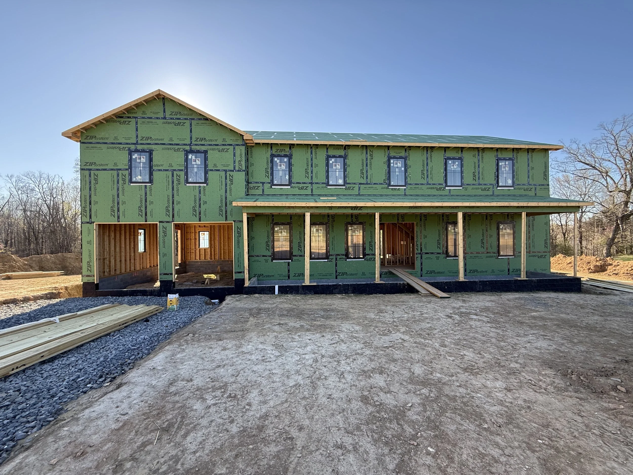 A house under construction with green sheathing, multiple windows, and a wooden roof structure. The site is outdoors with a clear blue sky and some trees in the background.