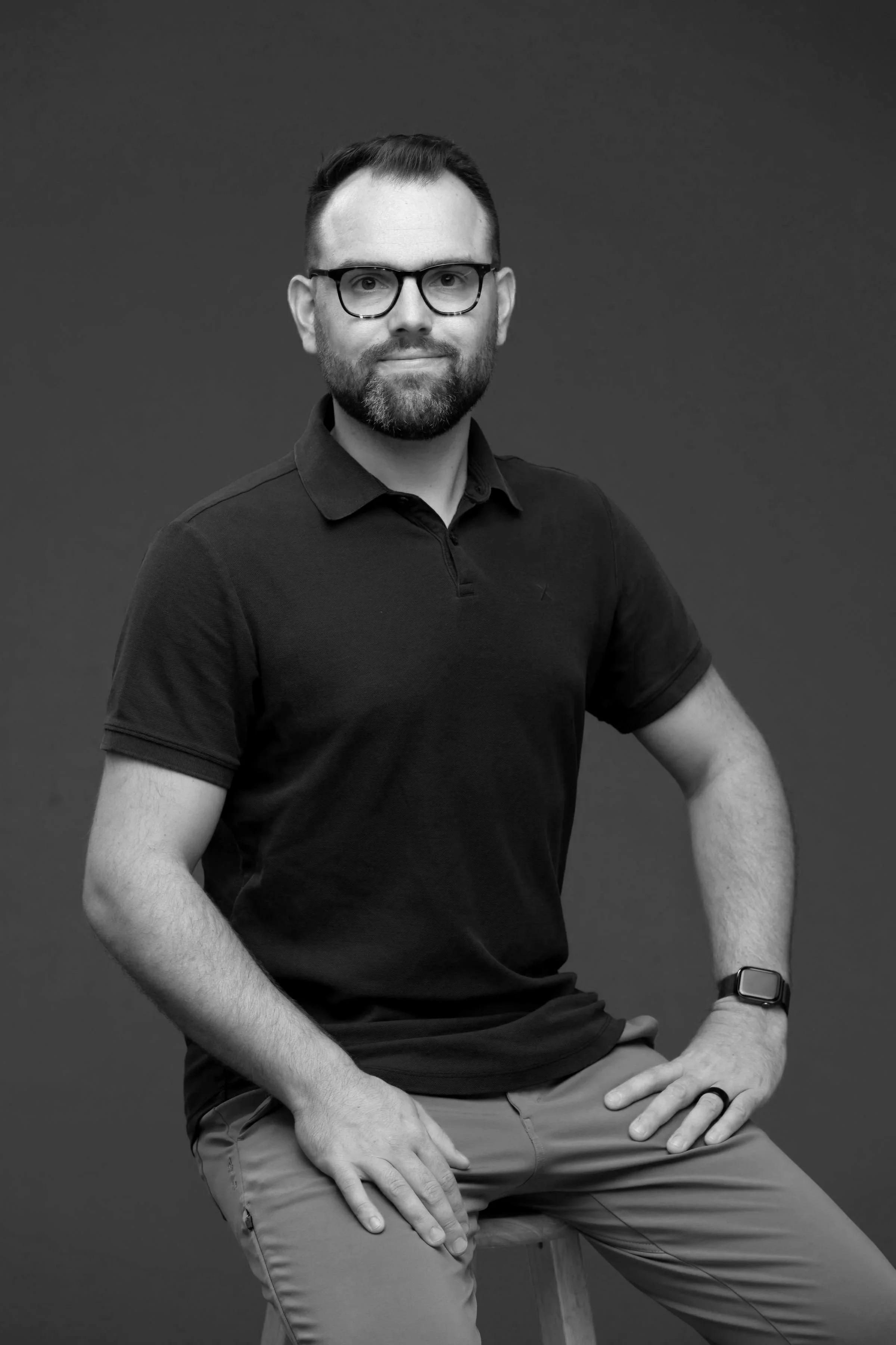 Black and white portrait of a man with glasses, beard, and short hair, wearing a polo shirt and khakis, seated with one hand on his knee, against a plain dark background.