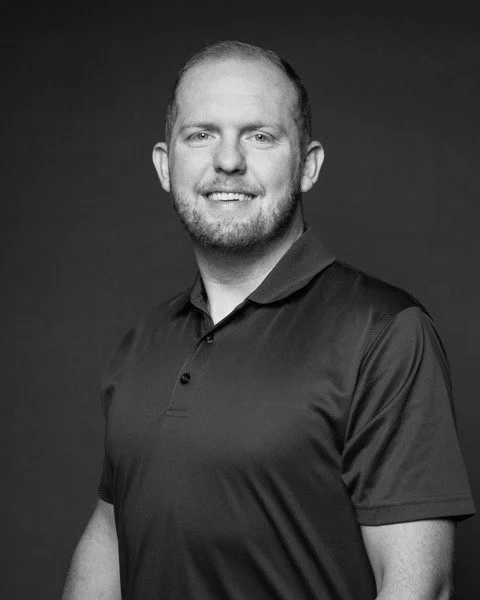 Black and white portrait of a smiling man with short hair and a beard, wearing a collared polo shirt, against a dark background.