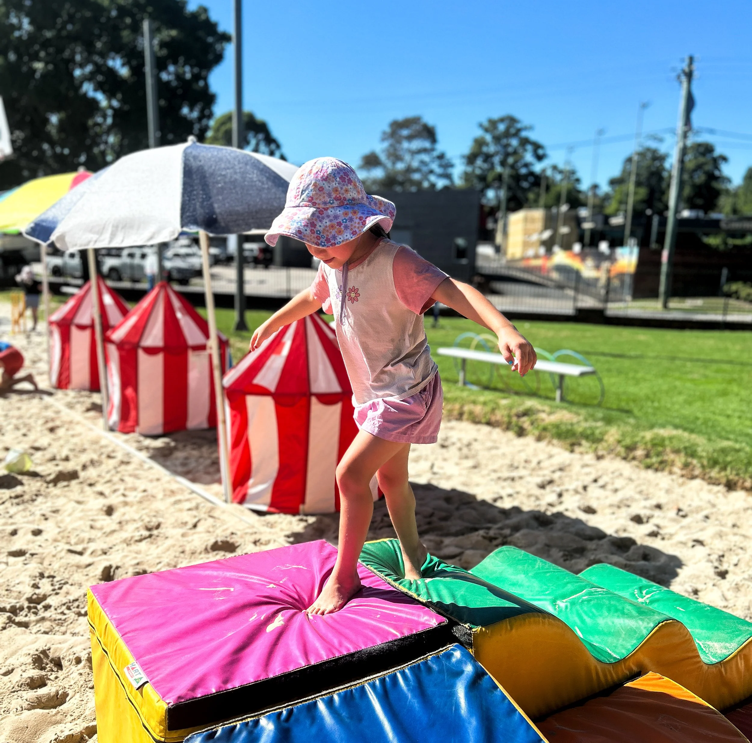 A young girl wearing a floral sunhat and pink shorts is jumping on a colorful inflatable bounce pad at a park or playground on a sunny day.